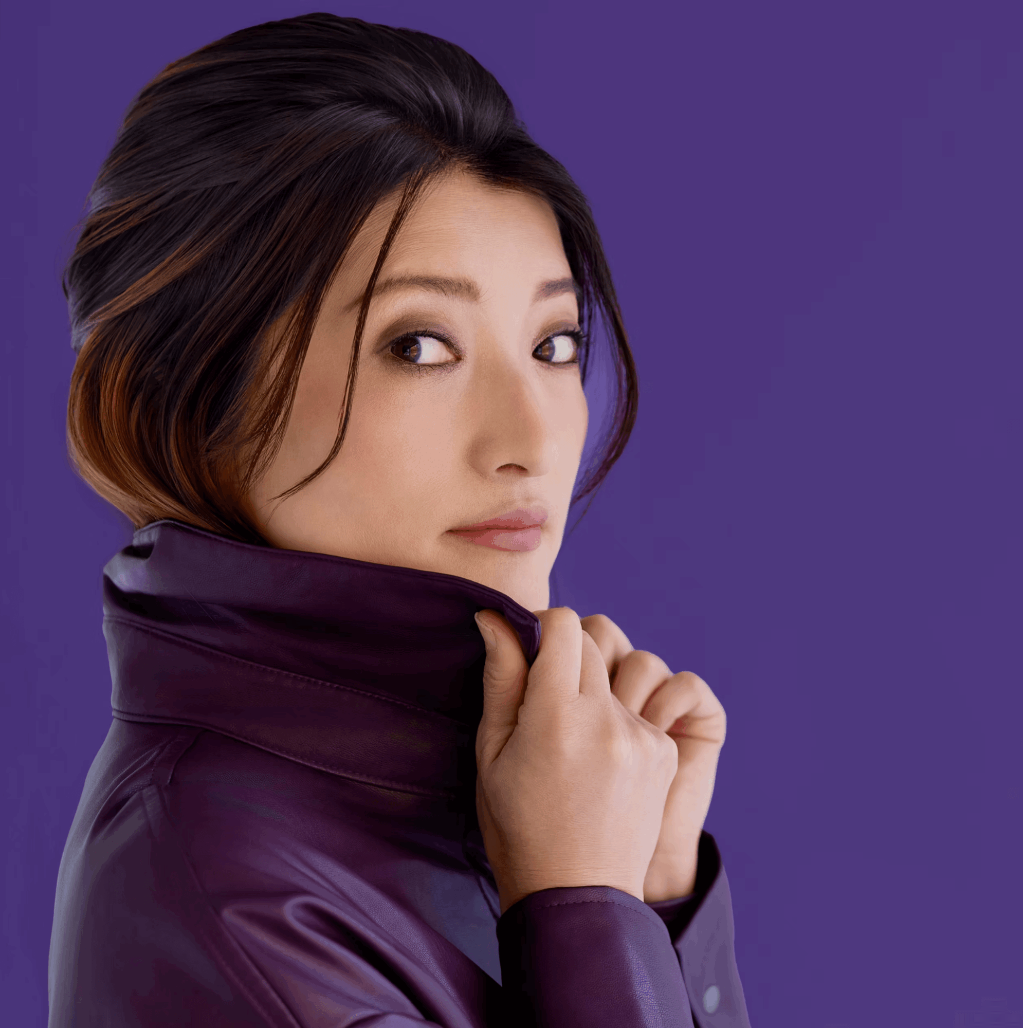 Woman with dark hair and makeup, wearing a purple jacket, looking over her shoulder against a purple background.