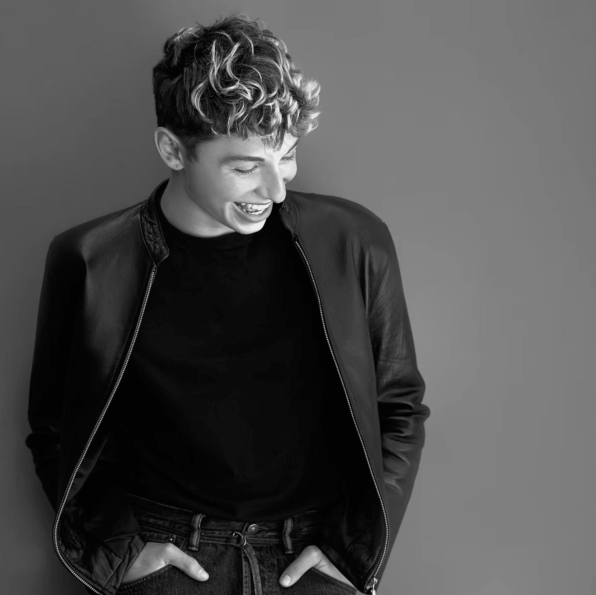 Young person with curly hair smiling, wearing a black jacket and dark shirt, hands in pockets, against a plain background.