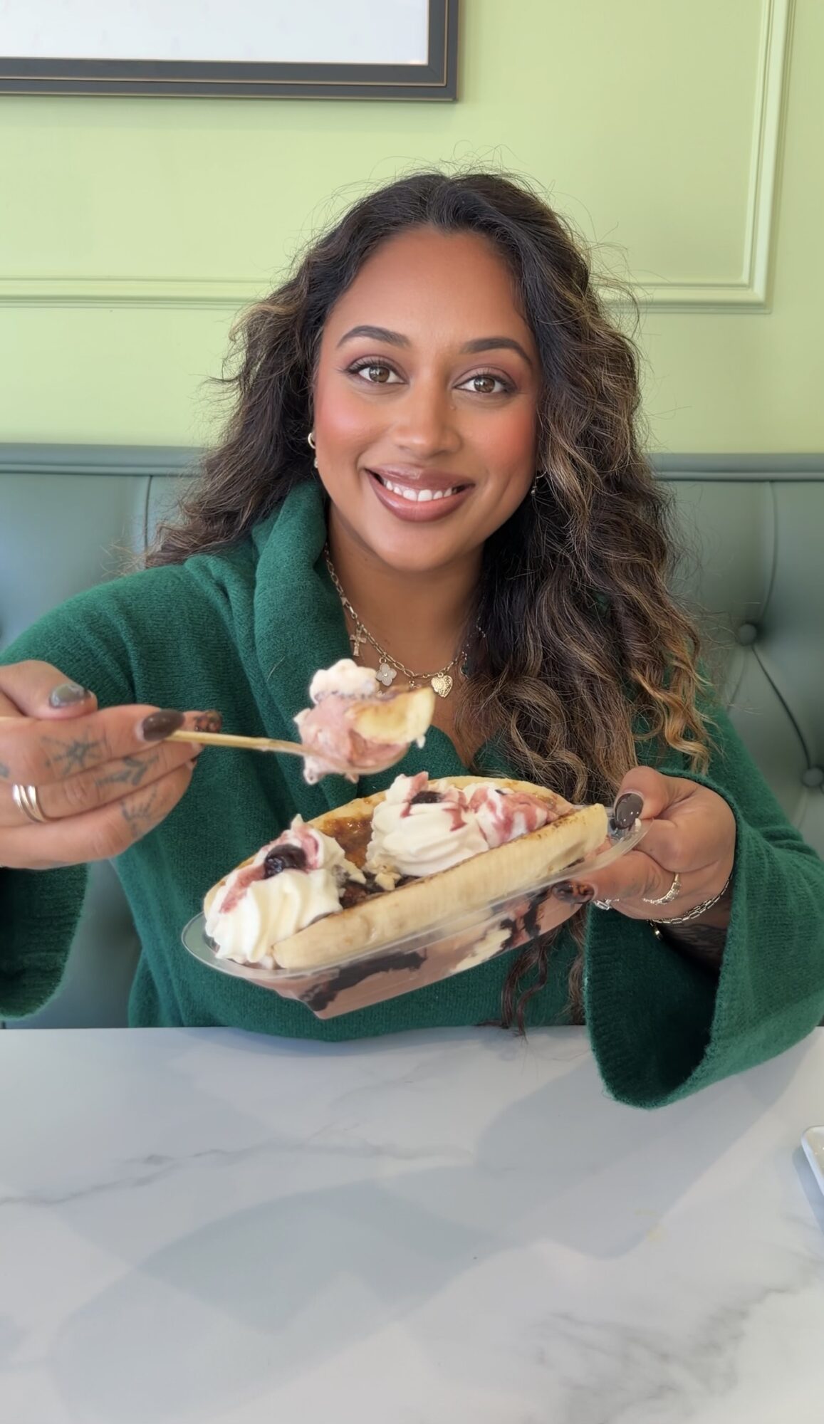 Woman with curly hair smiling, holding a bowl of ice cream with a spoon, sitting at a table in a restaurant.