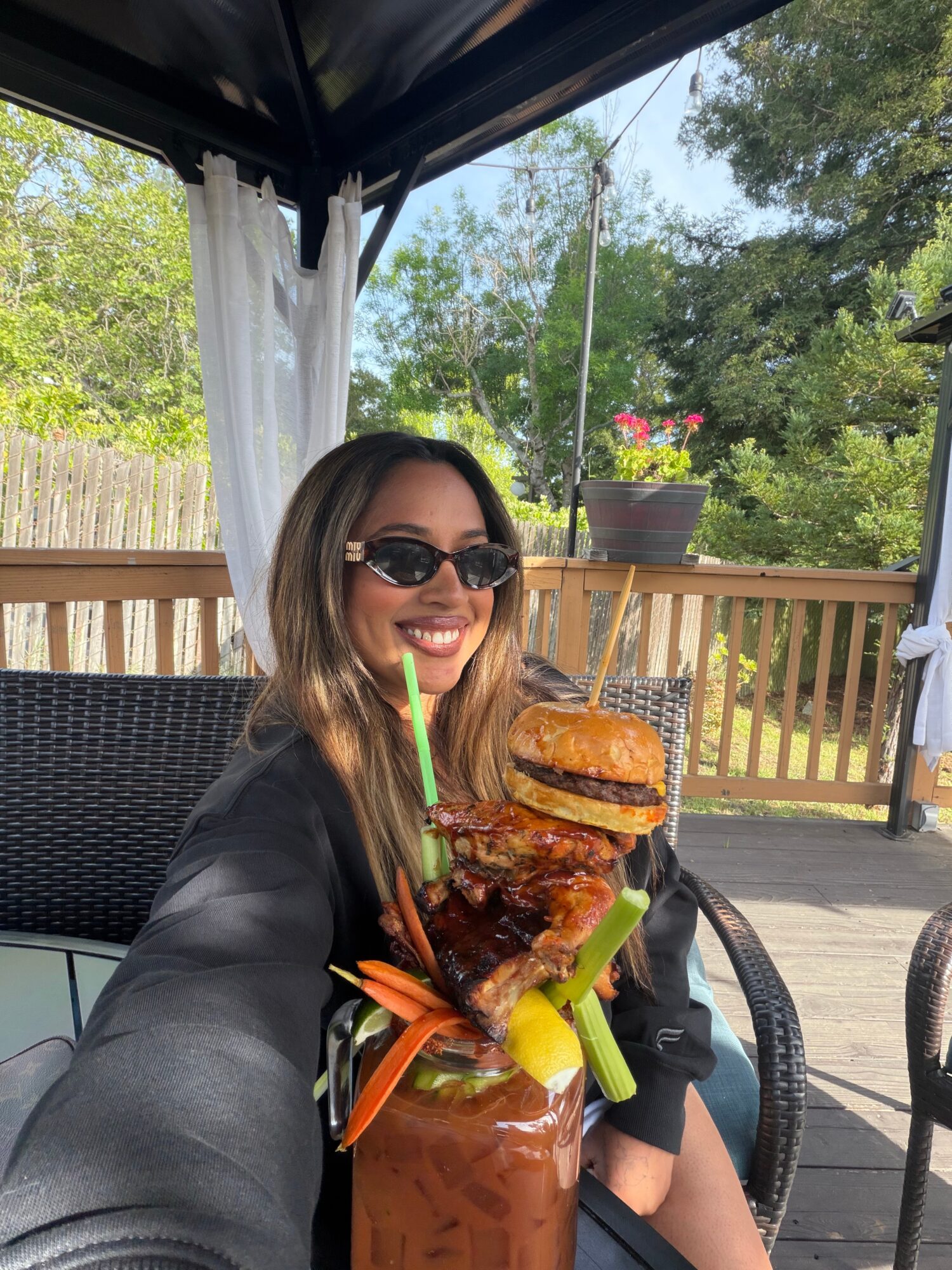 Woman with sunglasses smiling outdoors holding a large burger and a drink with vegetables, on a patio with trees and a fence.