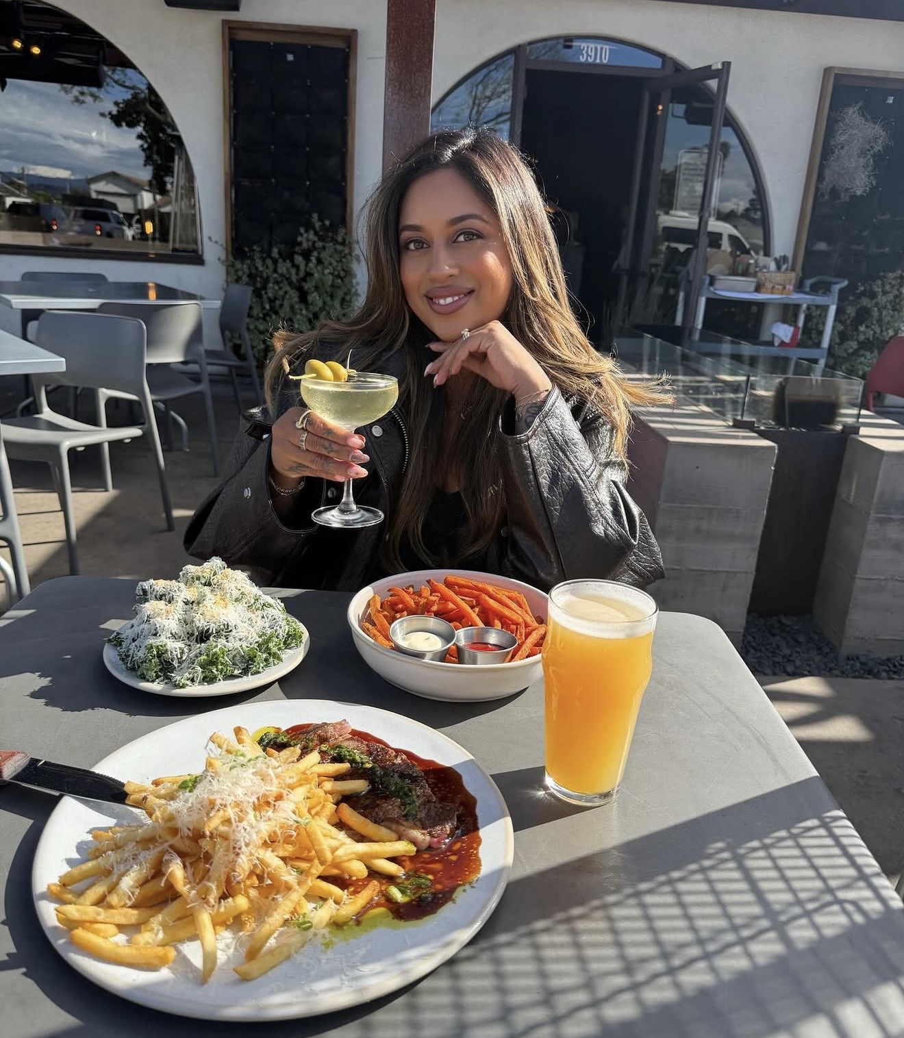 Woman sitting outdoors at a table with food and drinks, smiling, holding a cocktail with lemon, sunny day.