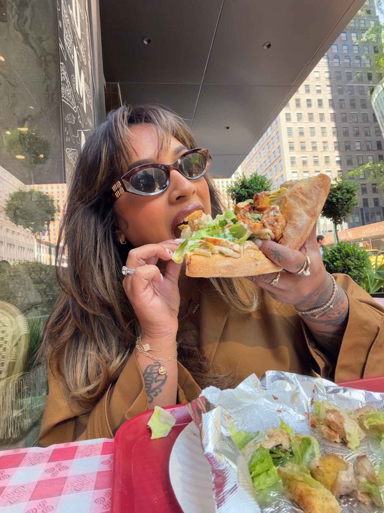 Woman with sunglasses eating a sandwich outdoors in an urban setting, with buildings and greenery in the background.