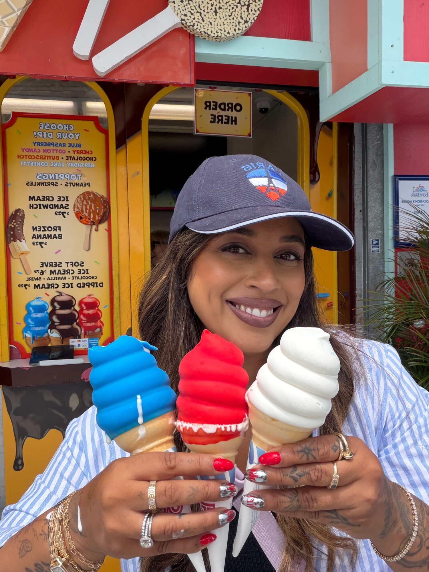 Woman smiling holding three ice cream cones in front of colorful storefront, wearing a cap and striped shirt.