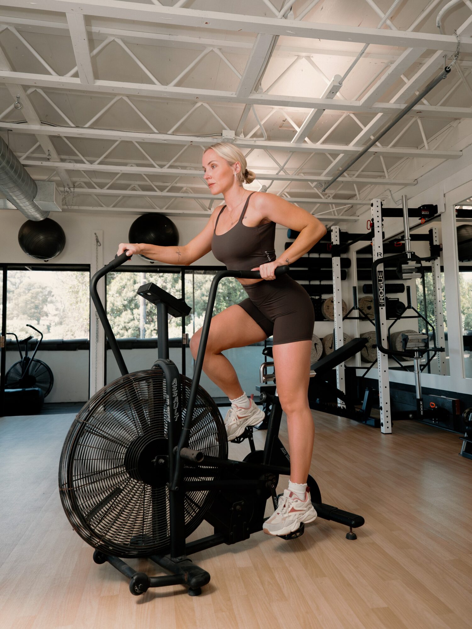 Woman using an air bike in a gym with weight racks and large windows, exercising with one foot on the pedal.