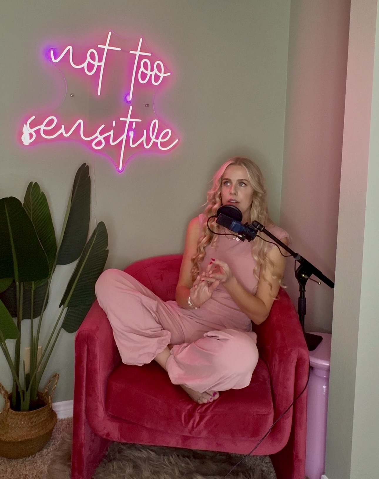 Woman with curly blonde hair sitting cross-legged on a pink velvet chair, holding a drink, with a neon sign reading 'not too sensitive' behind her.