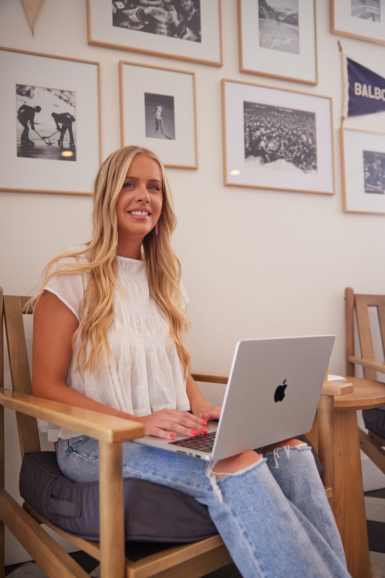 Woman with long blonde hair sitting at a wooden table with a laptop, smiling, in a room with framed pictures on the wall.