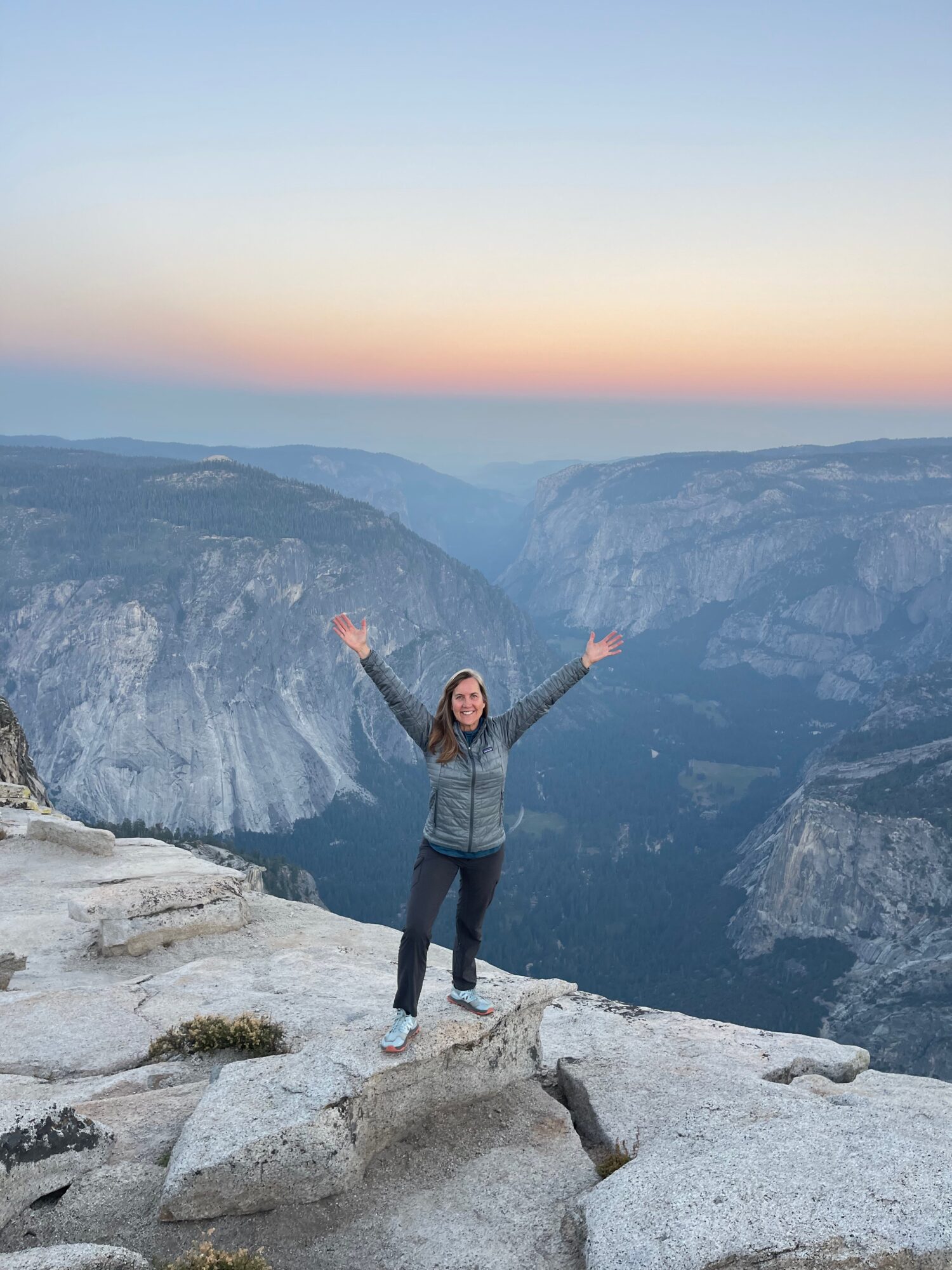 Person standing on rocks with arms raised, overlooking a canyon at sunset or sunrise, sky with gradient colors.