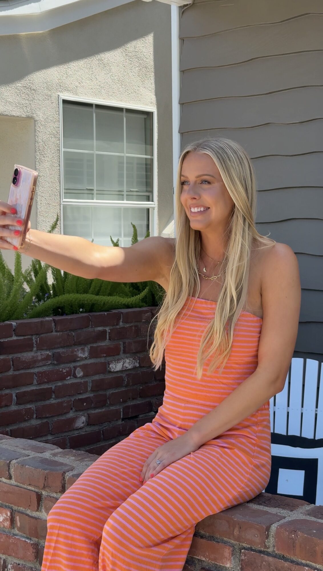 Woman with long blonde hair taking a selfie outdoors, sitting on a brick ledge, smiling, wearing a striped orange outfit.