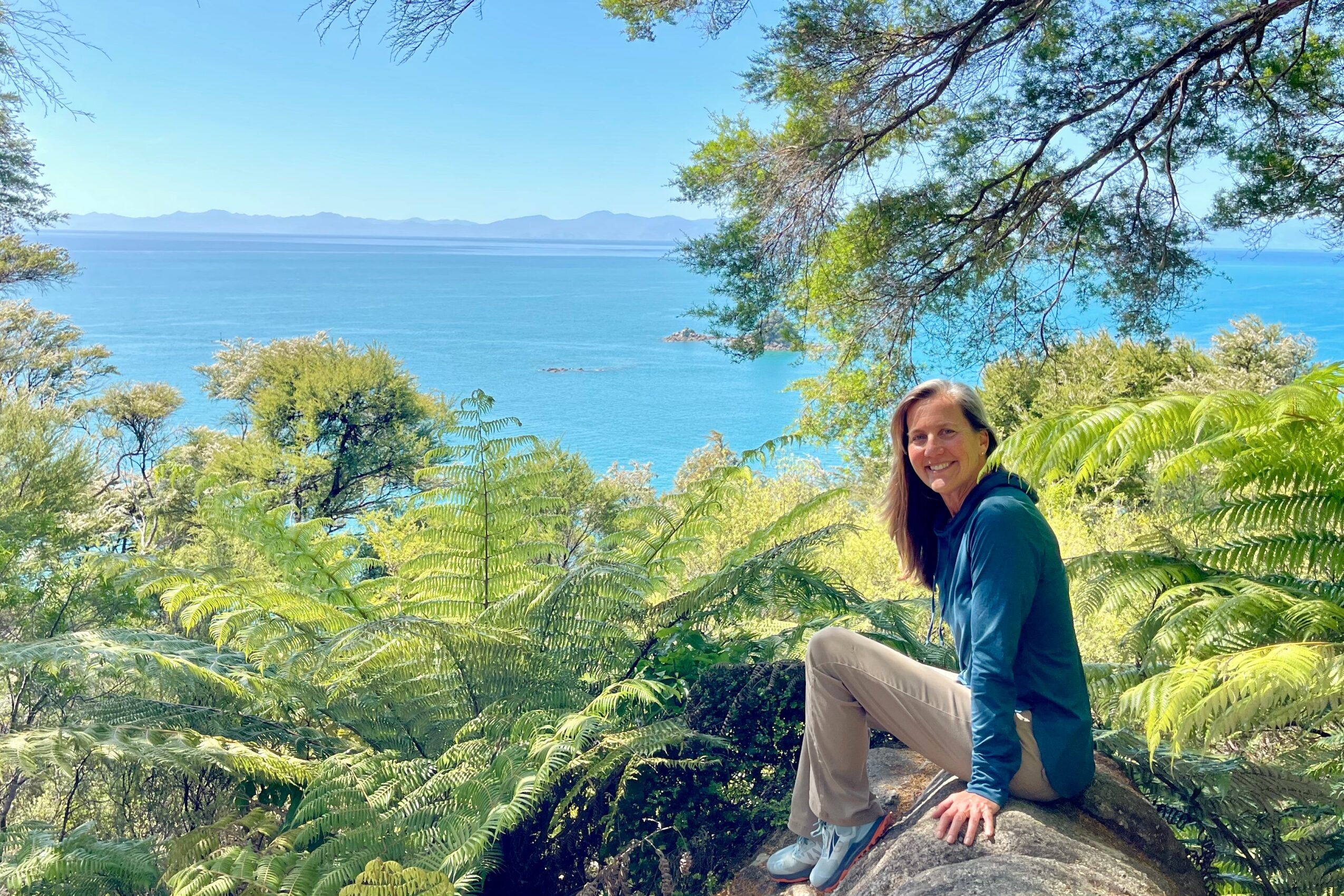 Woman sitting on a rock surrounded by lush greenery with a large body of water and mountains in the background.