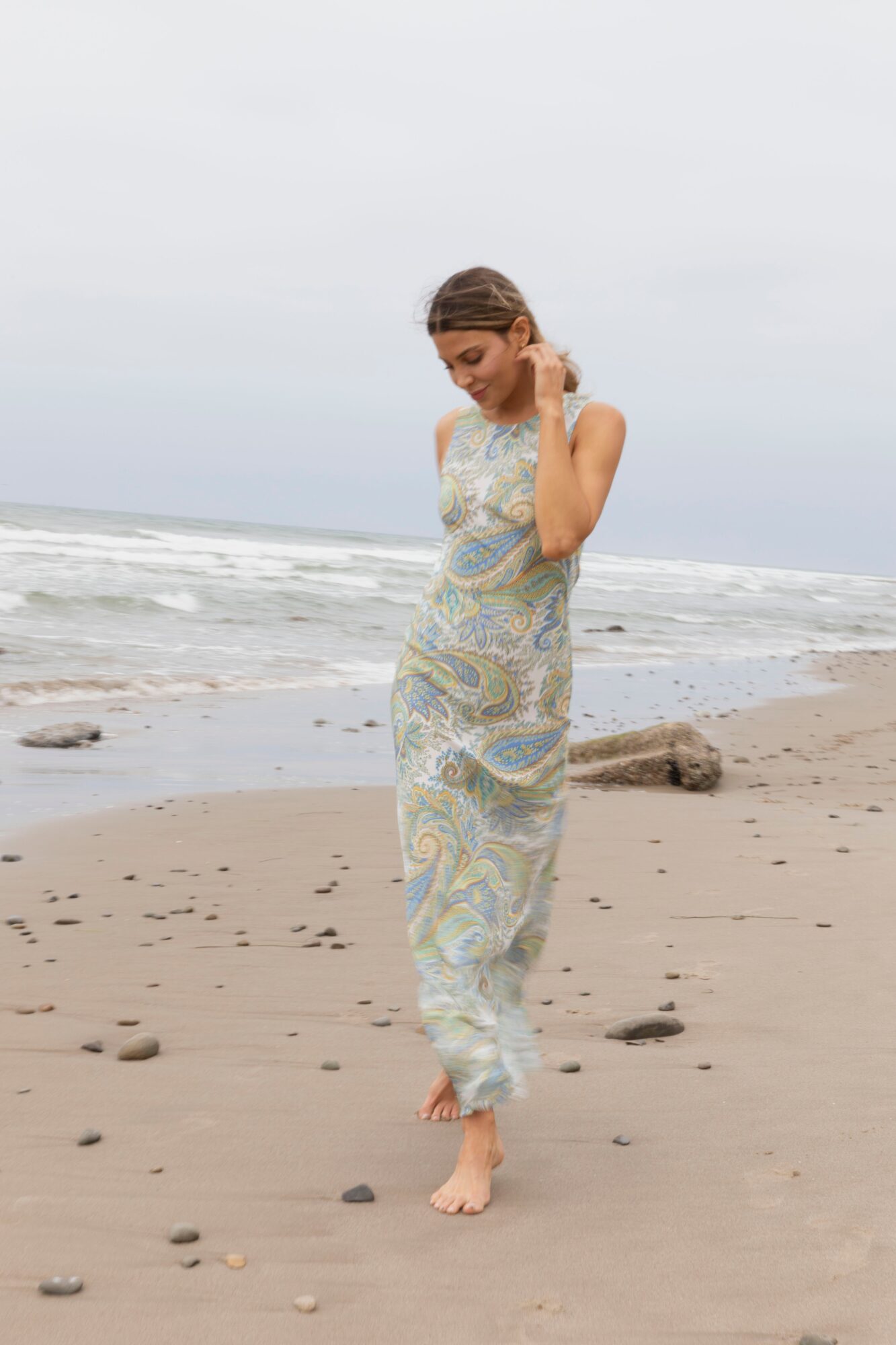 Woman in a patterned dress walking barefoot on the beach, looking down, with ocean waves in the background.