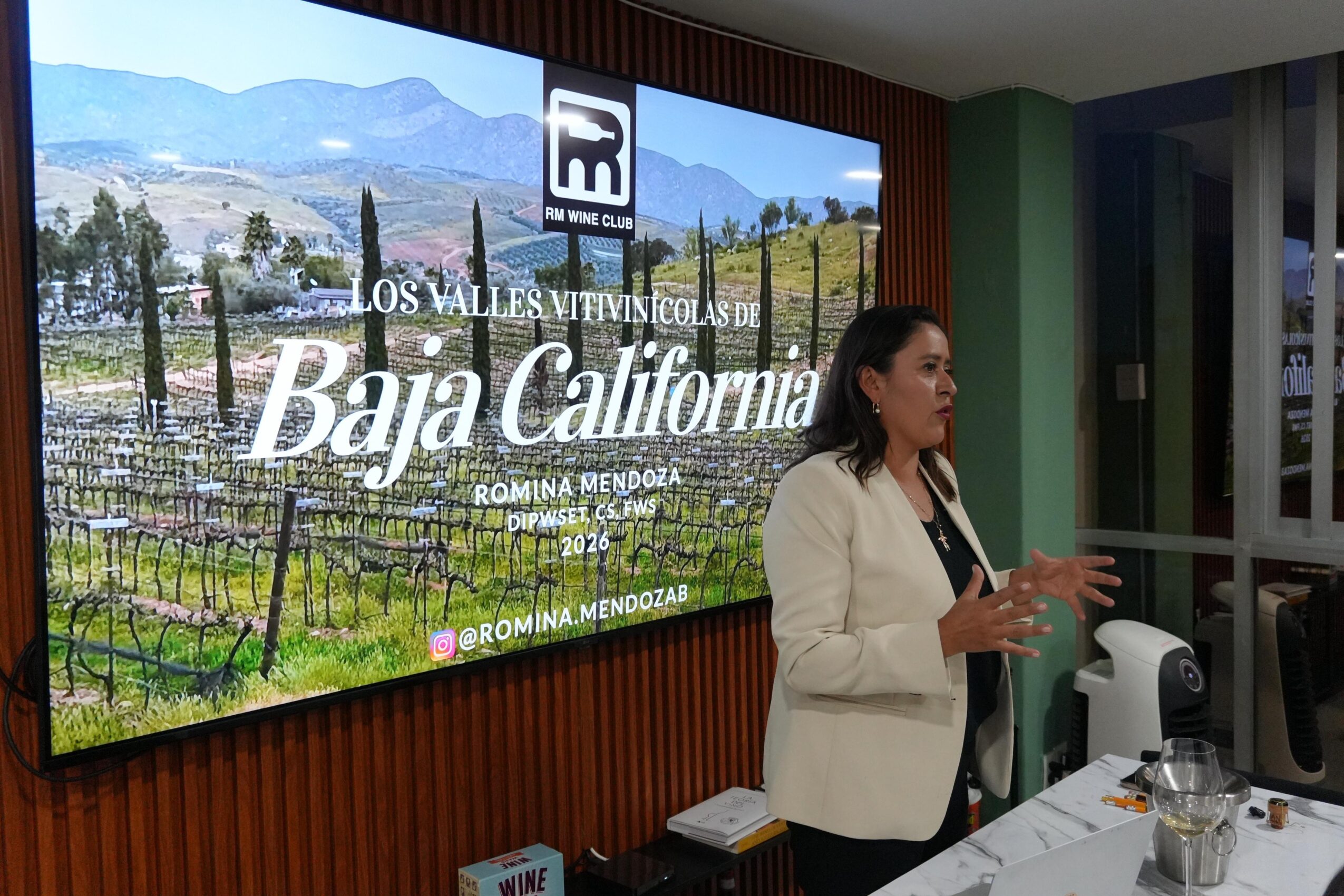 Woman in white blazer speaking in front of a large screen displaying Baja California landscape and text.