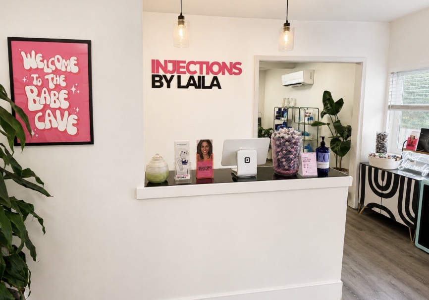 Reception area with a white counter, pink and black sign, framed pink poster, and decorative items, including plants and a window.
