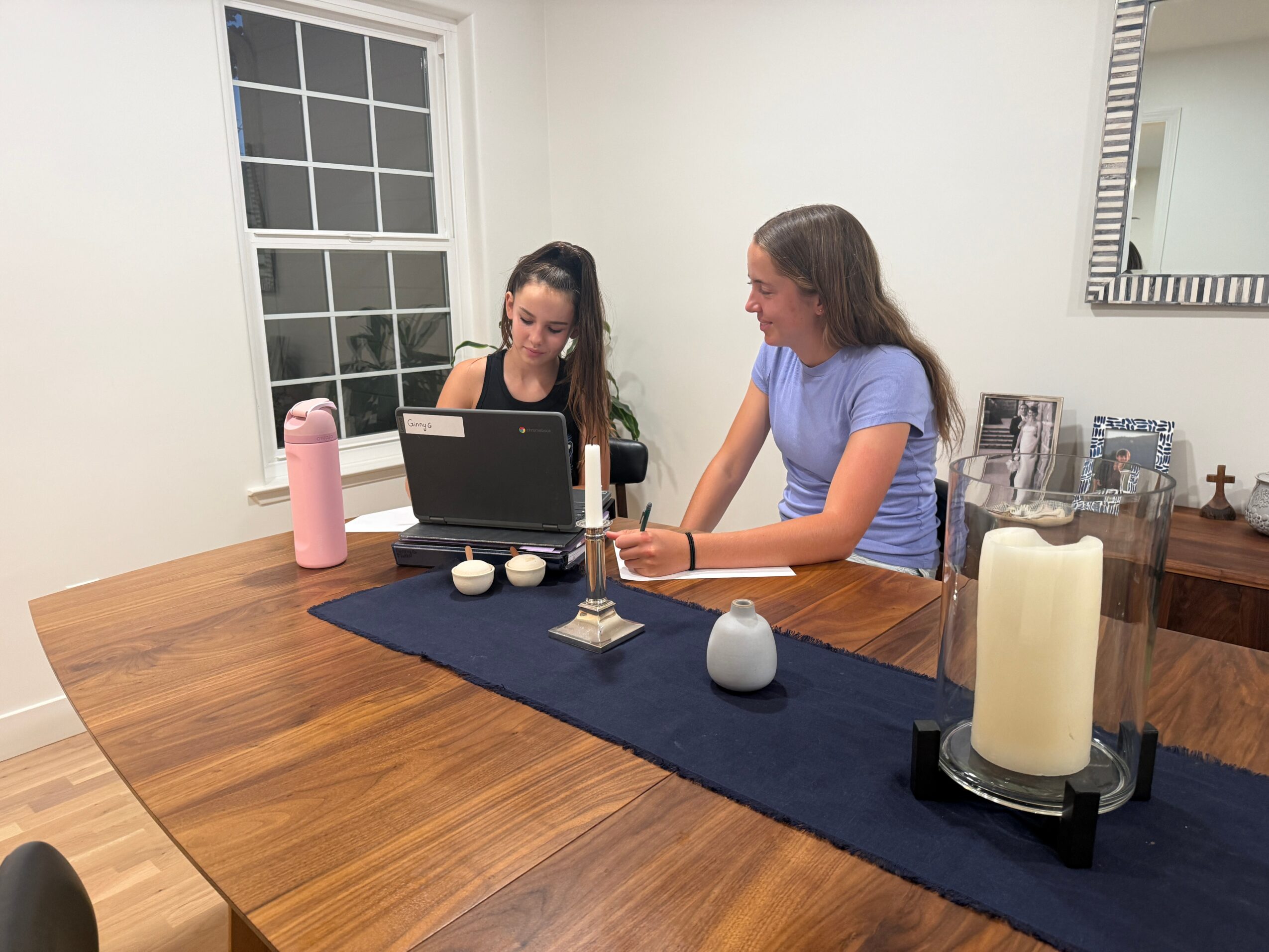 Two women sit at a wooden table with a laptop, candles, and decorative items in a bright room.