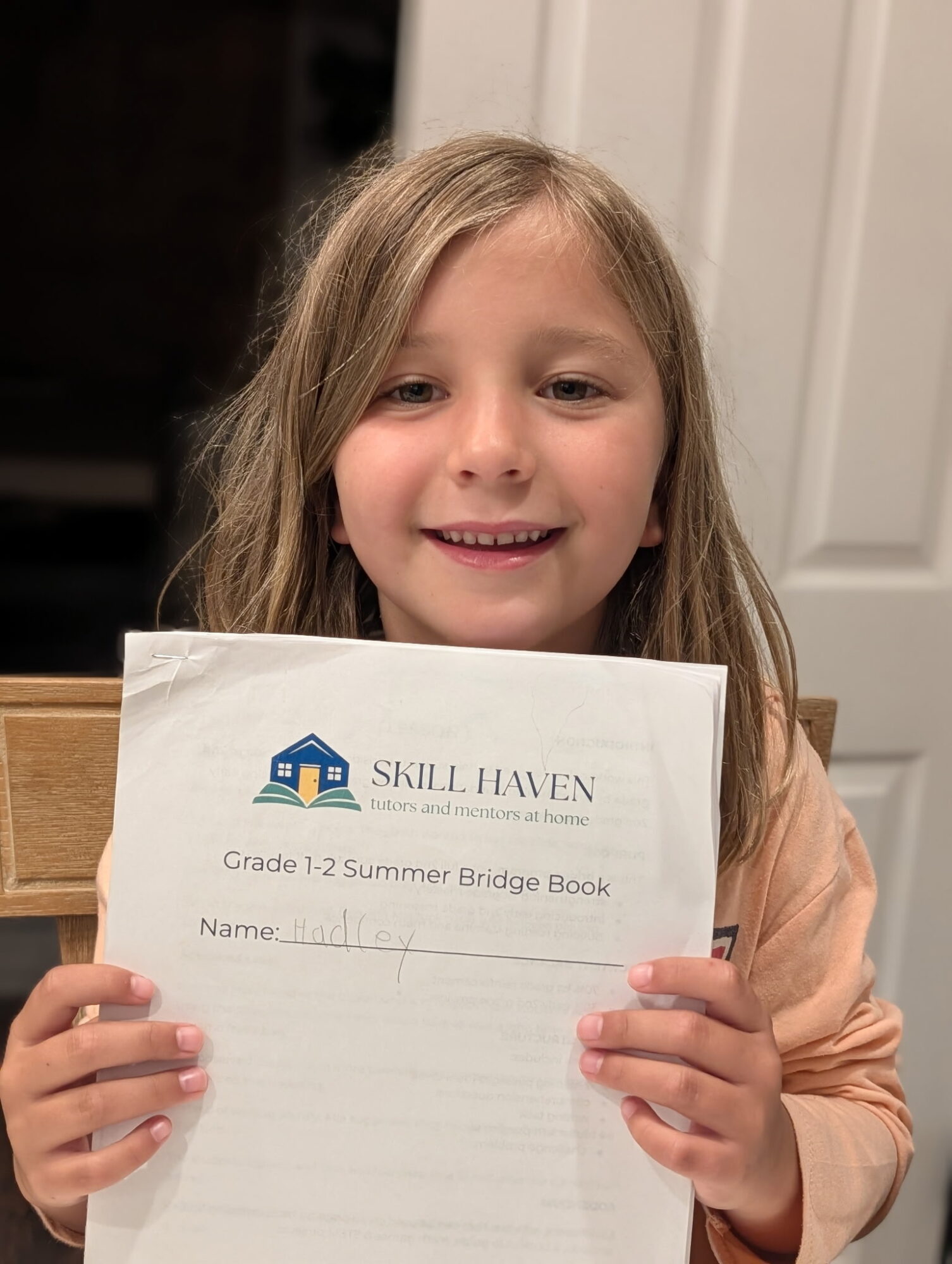 Young girl with long hair holding a Skill Haven summer bridge book, smiling, indoors.
