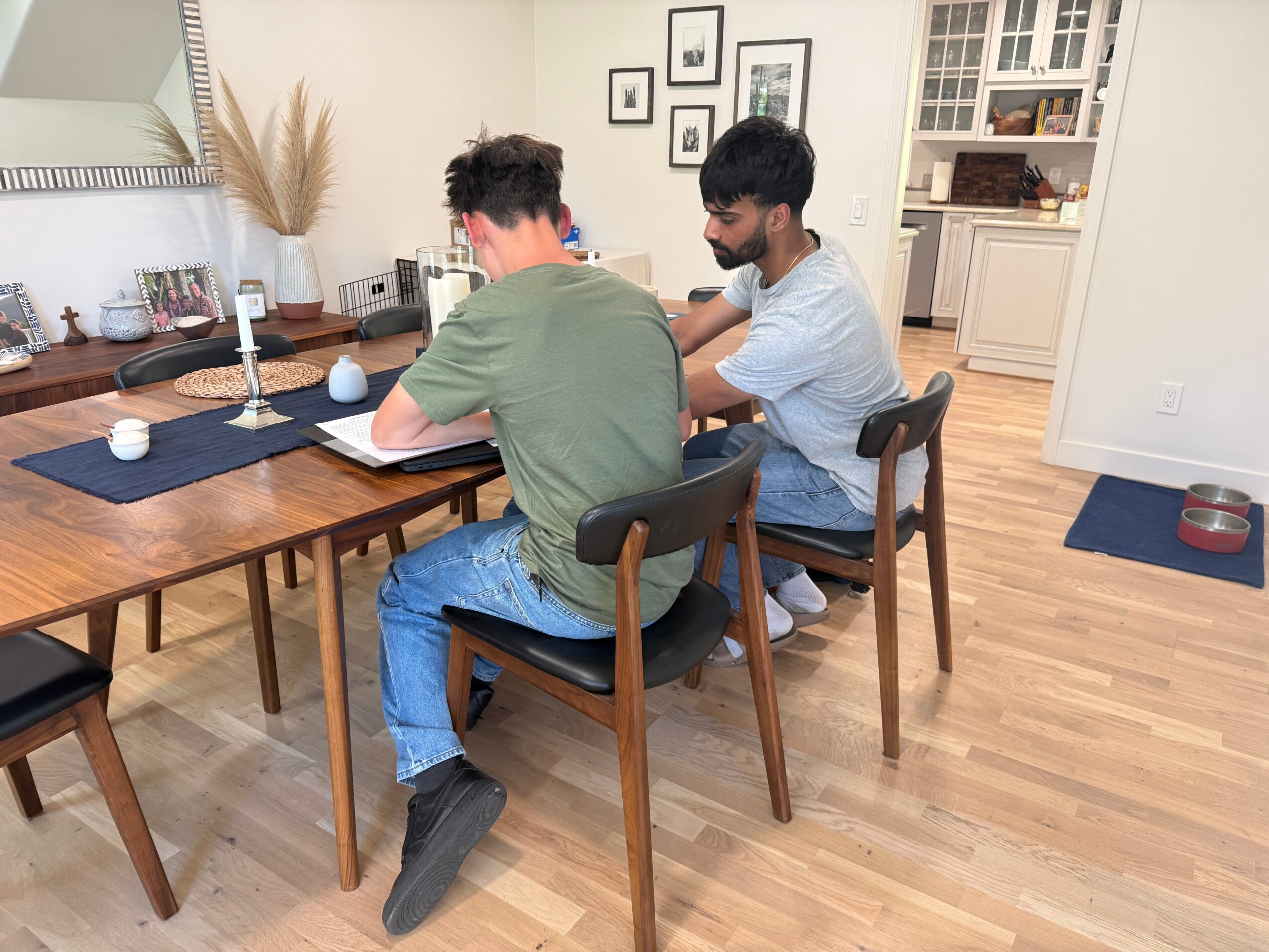 Two people sitting at a dining table in a bright kitchen, engaged in conversation or activity, with framed pictures on the wall.