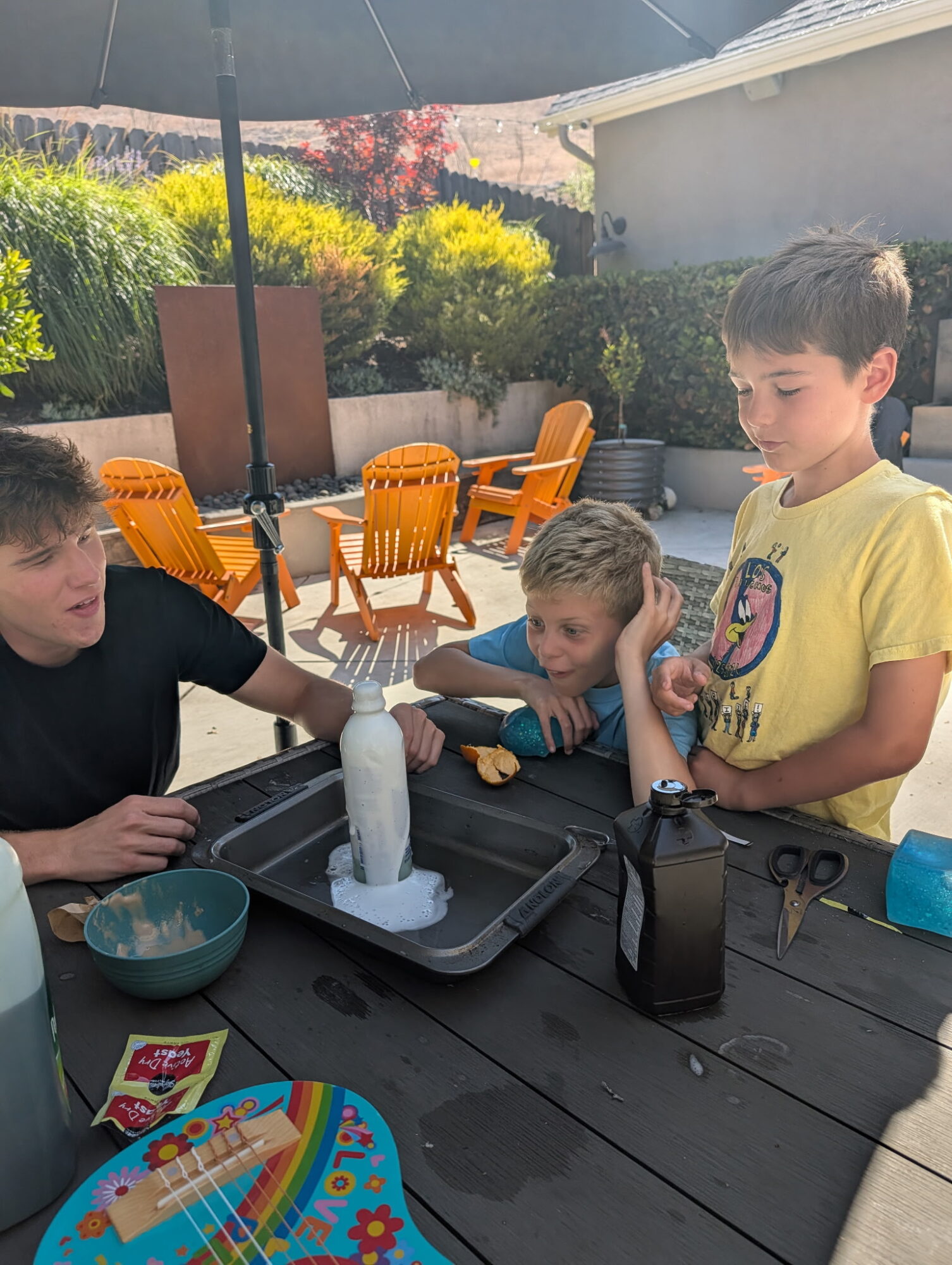Three children conduct a science experiment outdoors at a table with a bottle, tray, and supplies, under a large umbrella.