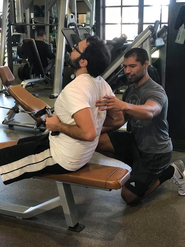 Person receiving a back massage from a trainer in a gym, seated on a massage bench, with gym equipment in background.