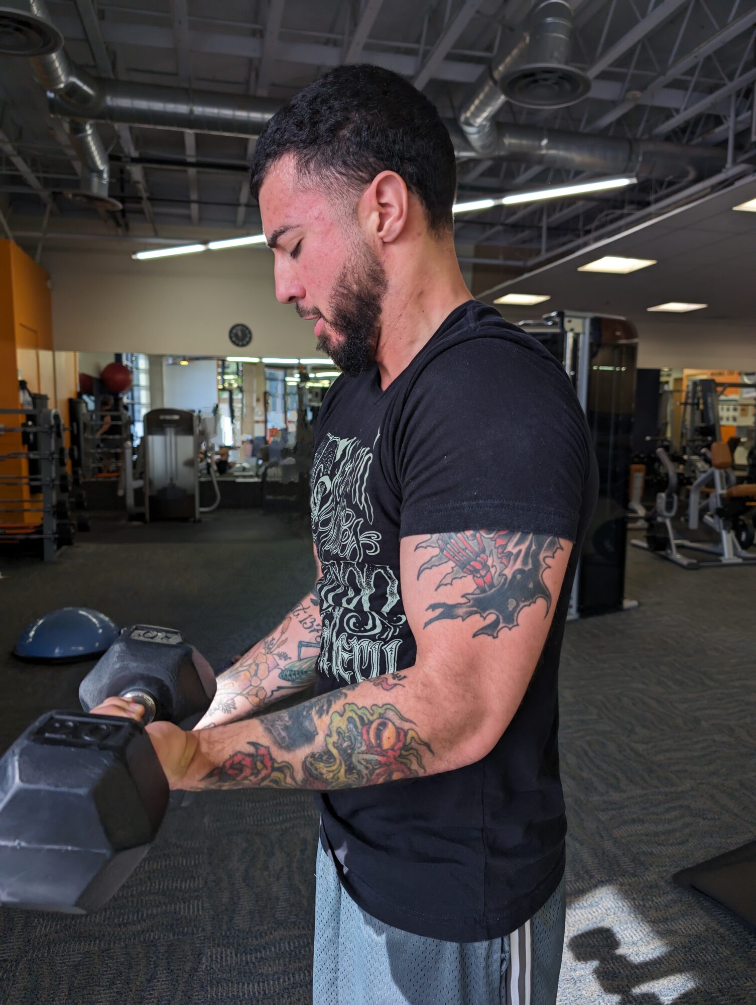 Man with tattoos on arms lifting a dumbbell in a gym, side view, focused expression.