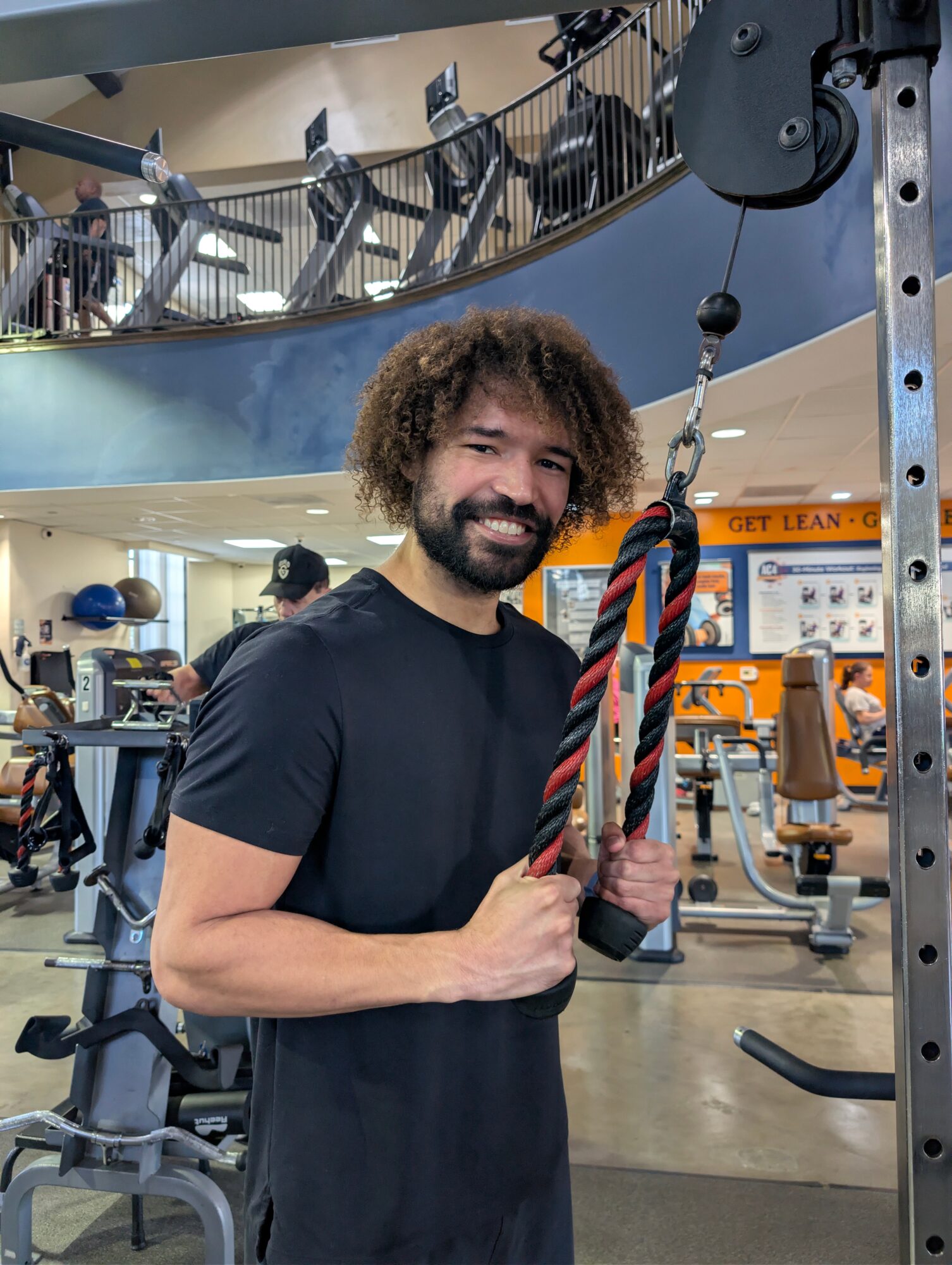 Man with curly hair and beard smiling at gym, holding a rope attachment, surrounded by exercise equipment.