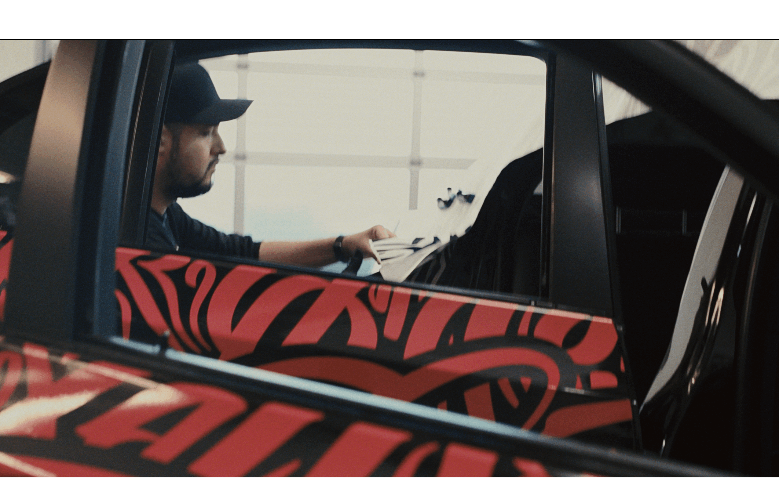Person with a beard and cap sitting in a vehicle, looking at a phone, with a red and black patterned seat cover visible.