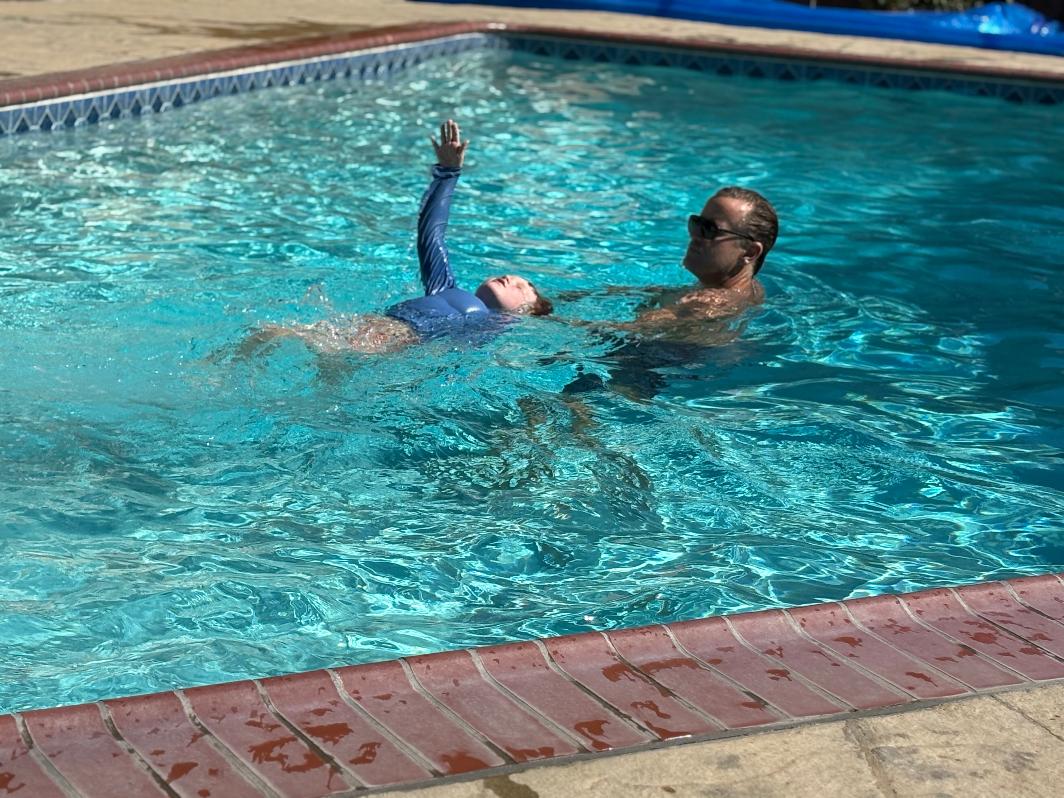 Child floating on back in swimming pool with adult holding and supporting them, pool edge visible, sunny day.