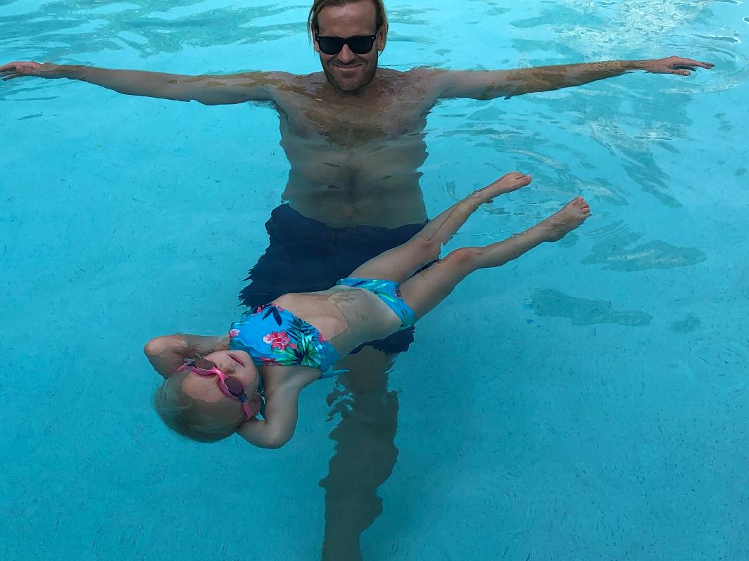 Man and girl floating in a swimming pool, man with arms outstretched, girl with sunglasses, both relaxed in water.