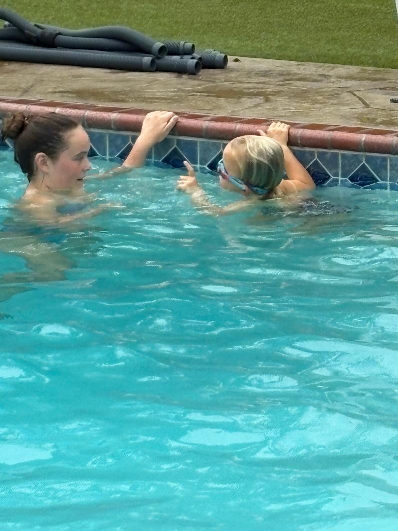 Two people in a swimming pool near the edge, one assisting the other with goggles, water visible, pool edge and grass in background.