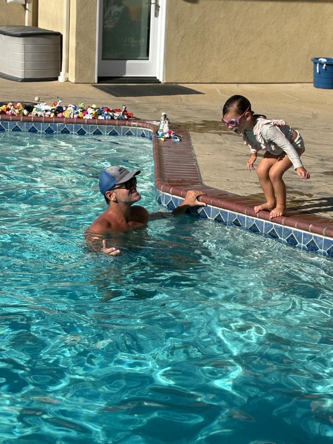 Child and adult near swimming pool edge, girl jumping into water, sunny outdoor setting, pool toys on deck, building in background
