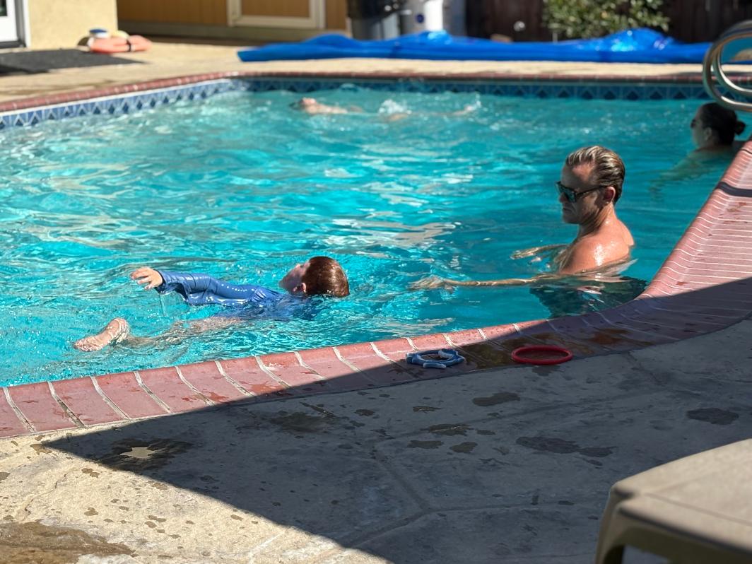 Child swimming in pool with adult supervising nearby, poolside area visible, sunlight reflecting on water.