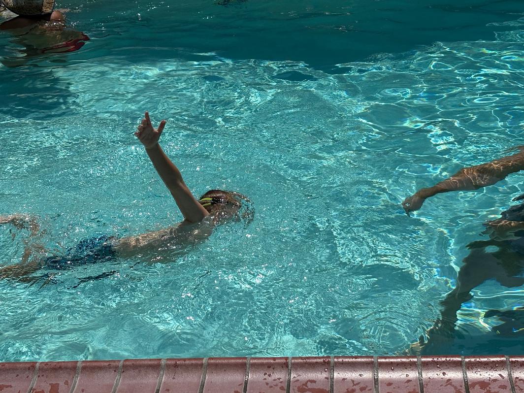 Child swimming in pool with arm raised, water ripples visible, pool edge at bottom, other swimmers partially visible