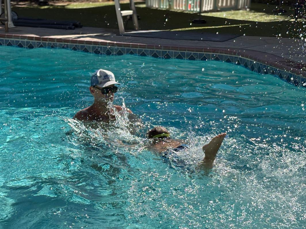 Person swimming in a pool with a cap, splashing water, and a sunny outdoor setting.