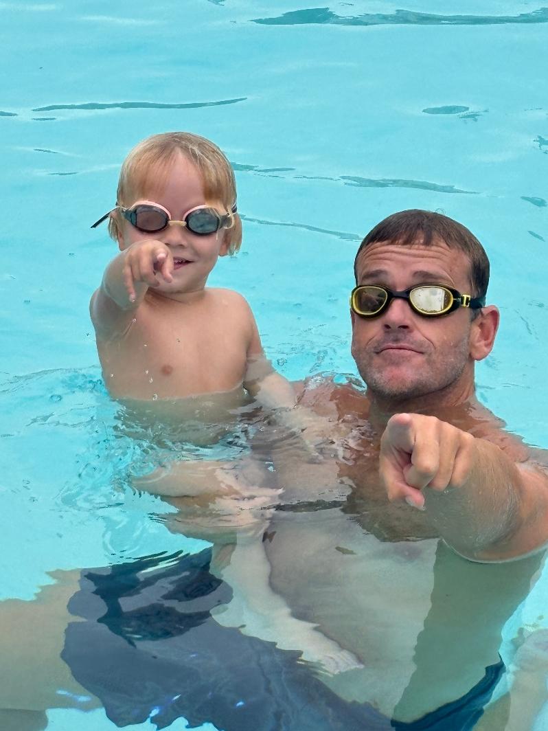 A man and a young girl wearing goggles in a swimming pool, pointing at the camera, smiling, water around them.