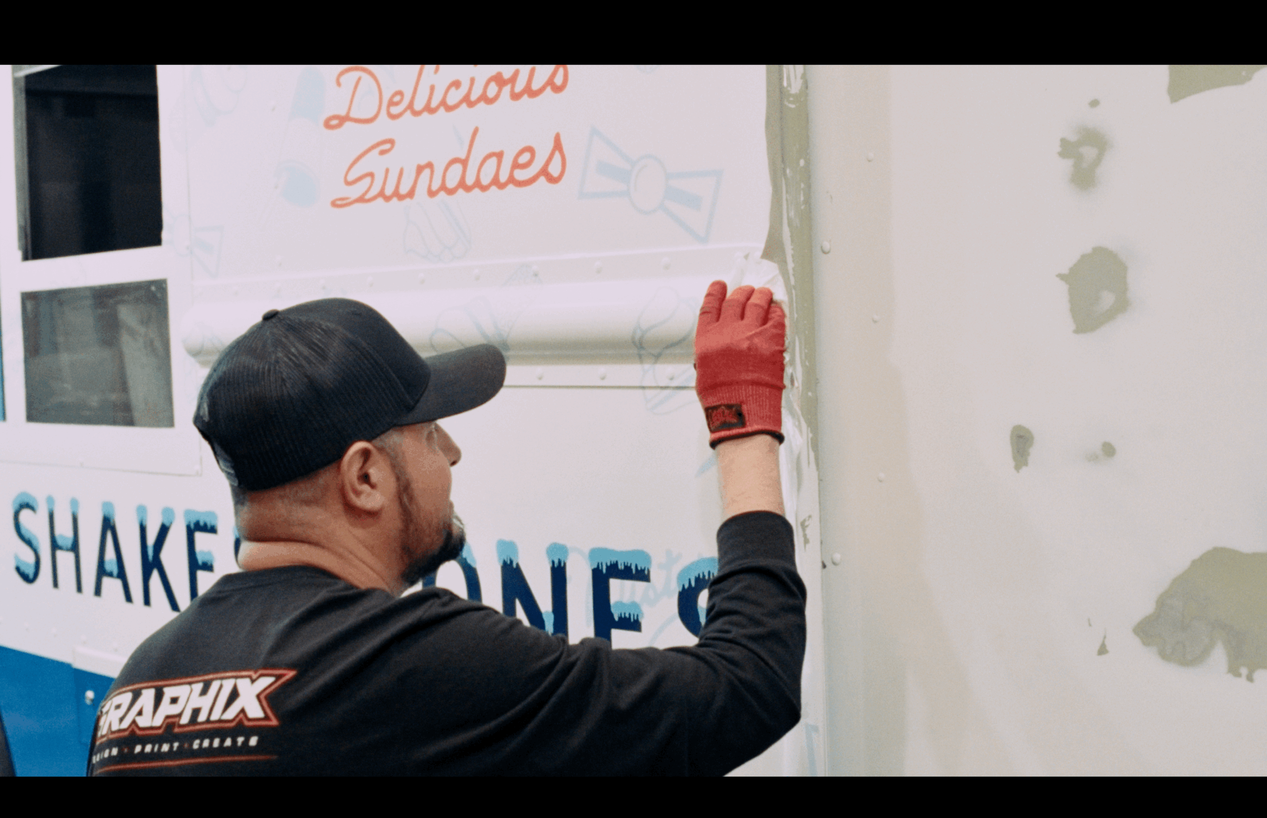 Person wearing a black cap and red glove writing on a wall, with a sign reading 'Delicious Sundaes' in the background.