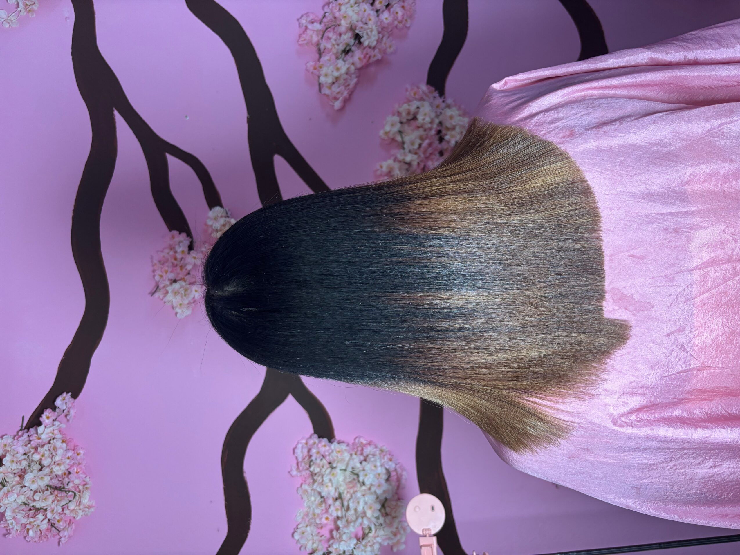 Person with long, straight hair wearing a pink satin garment, standing in front of a purple wall with black tree branches and pink flowers.