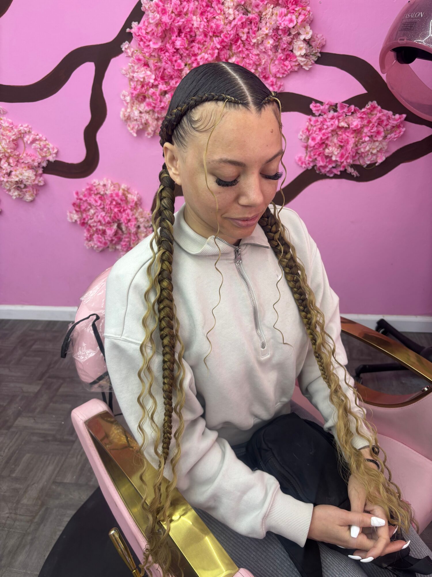 Young woman with long braided hair, wearing a white hoodie, sitting at a pink table with a pink background and cherry blossom decoration.