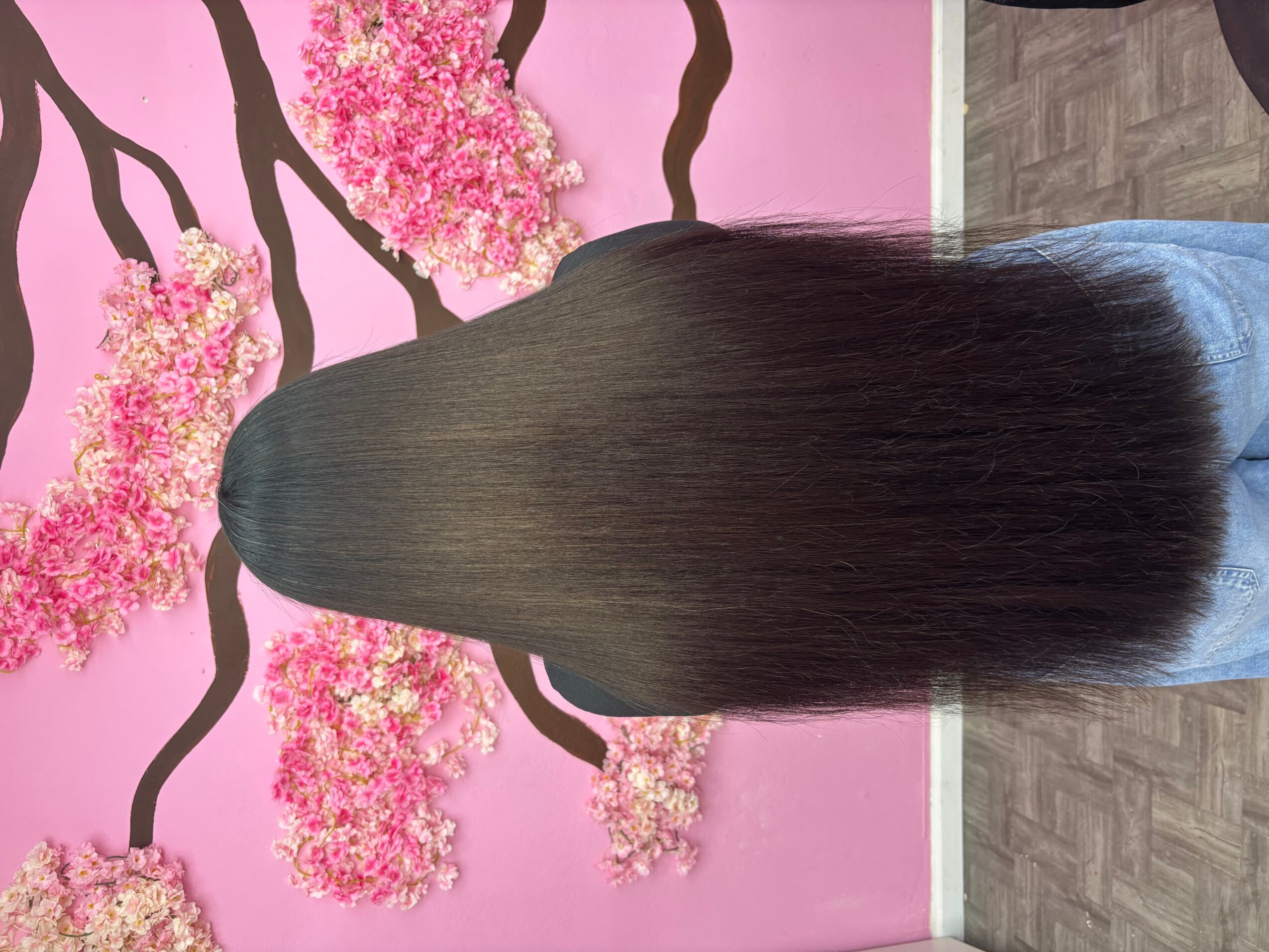 Person with long, straight, dark hair standing in front of pink floral wall, back view.
