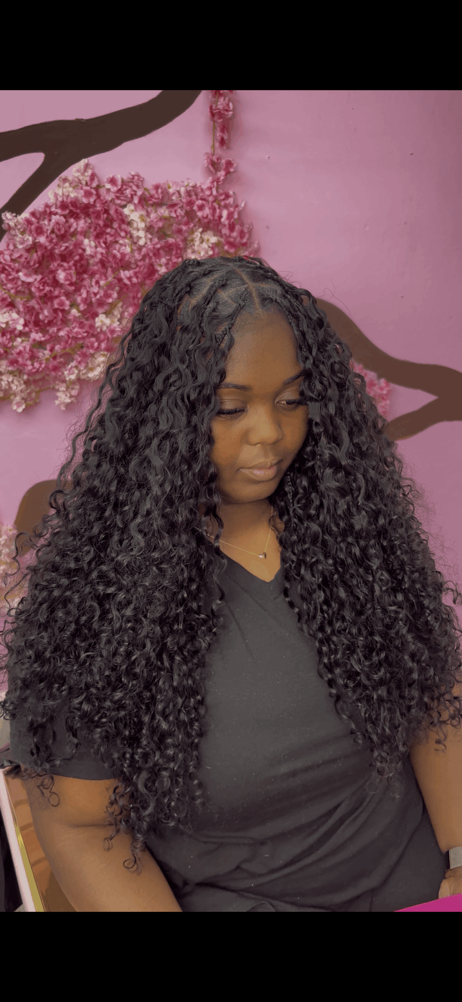 Young woman with long curly hair standing in front of pink wall with floral decoration, looking down.