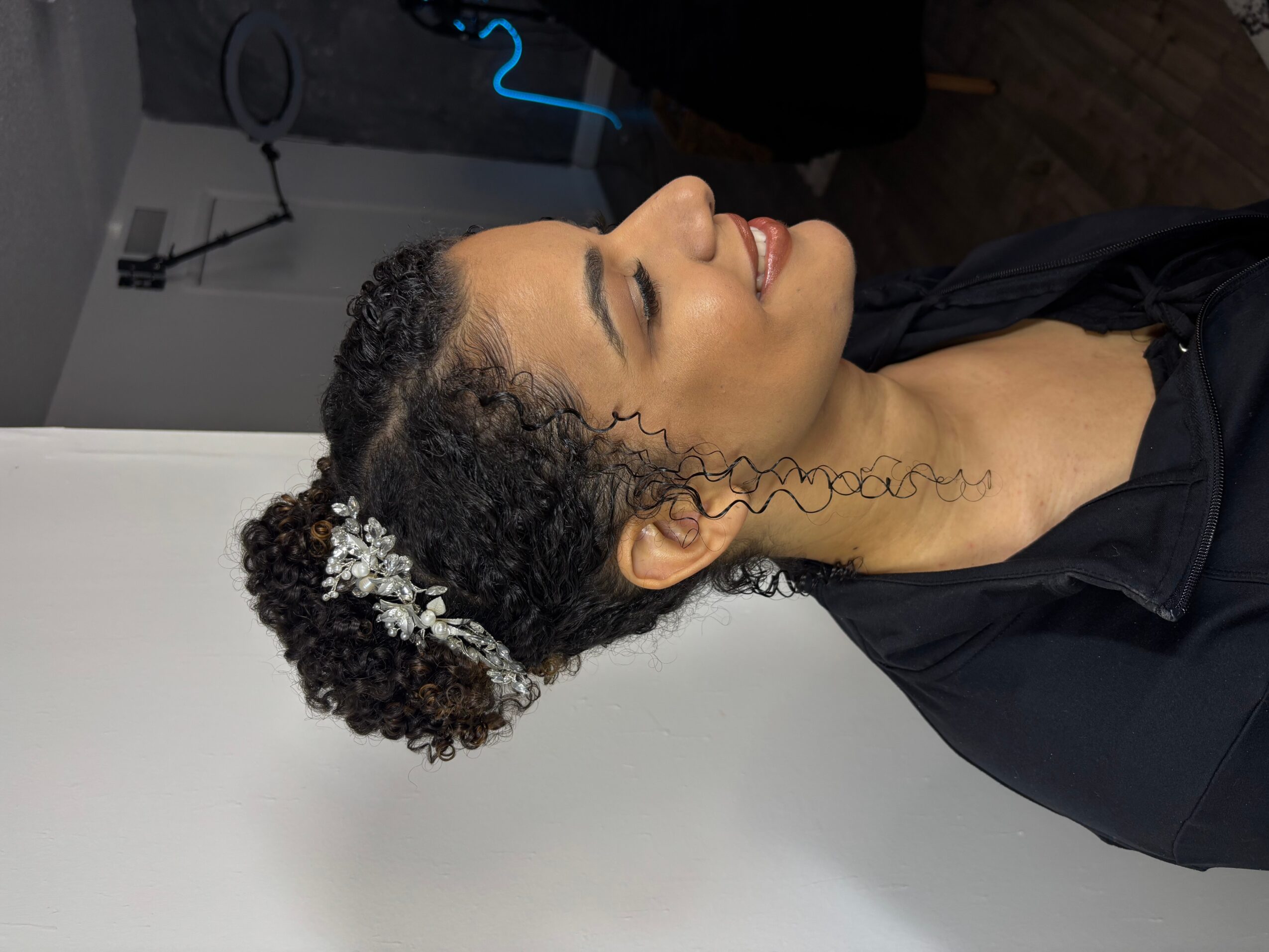 Side profile of a smiling woman with curly hair decorated with white flowers, wearing a black top.