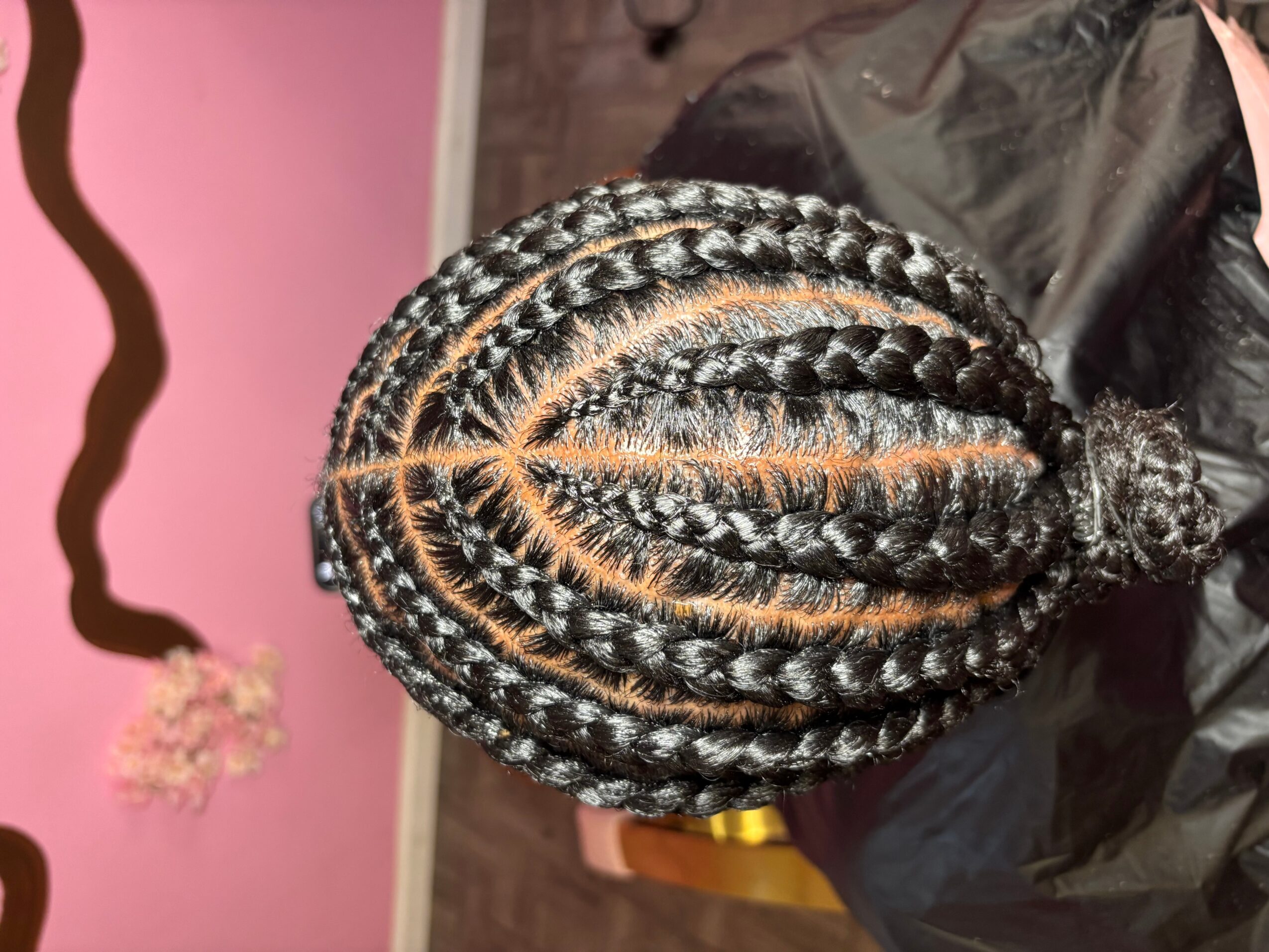 Top view of a person's head with braided hair, showing intricate cornrows, against a pink wall background.