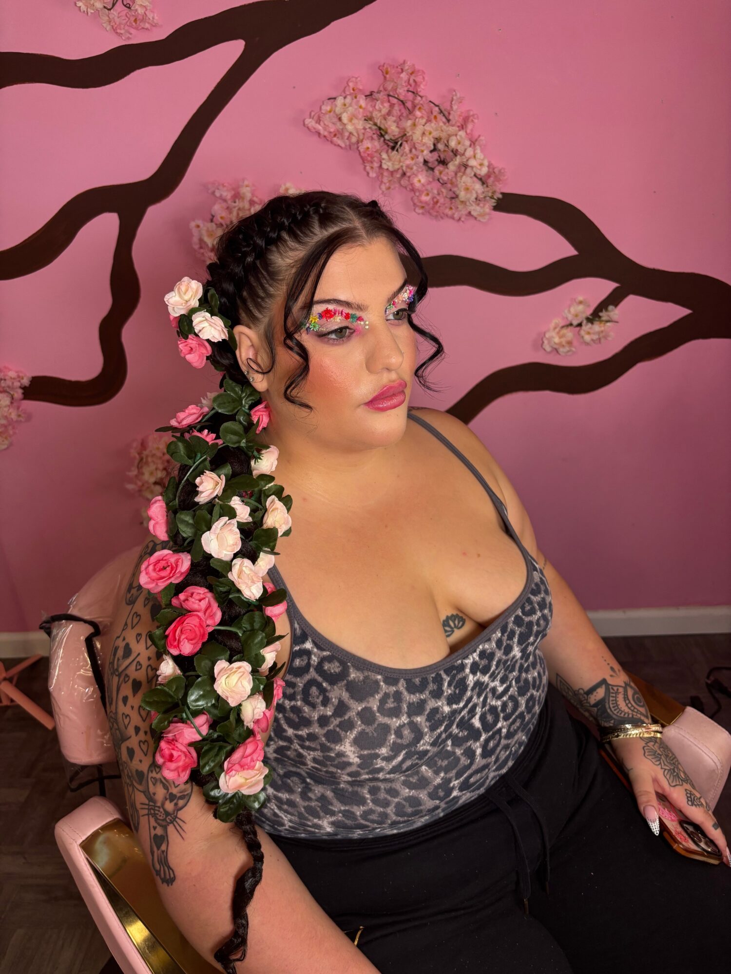 Woman with long braided hair decorated with flowers, lying on a pink wall with black branch designs, holding a phone.
