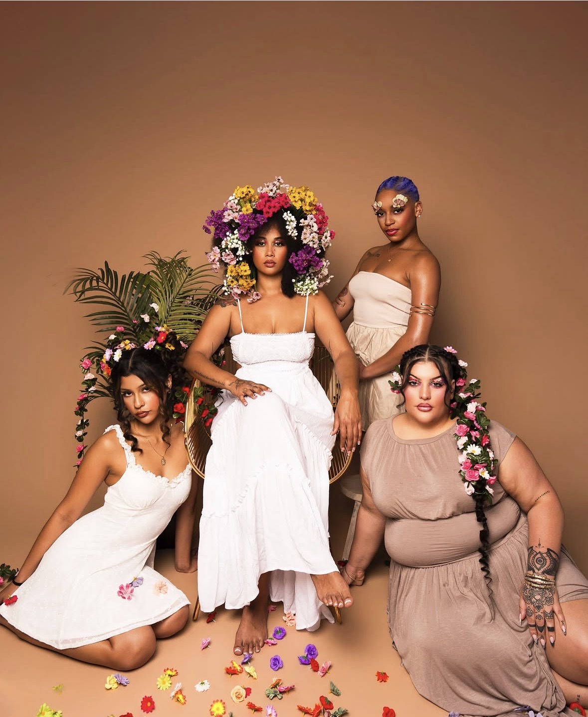 Four women in floral and white dresses pose against a plain background, with floral headpieces and accessories, looking at the camera.