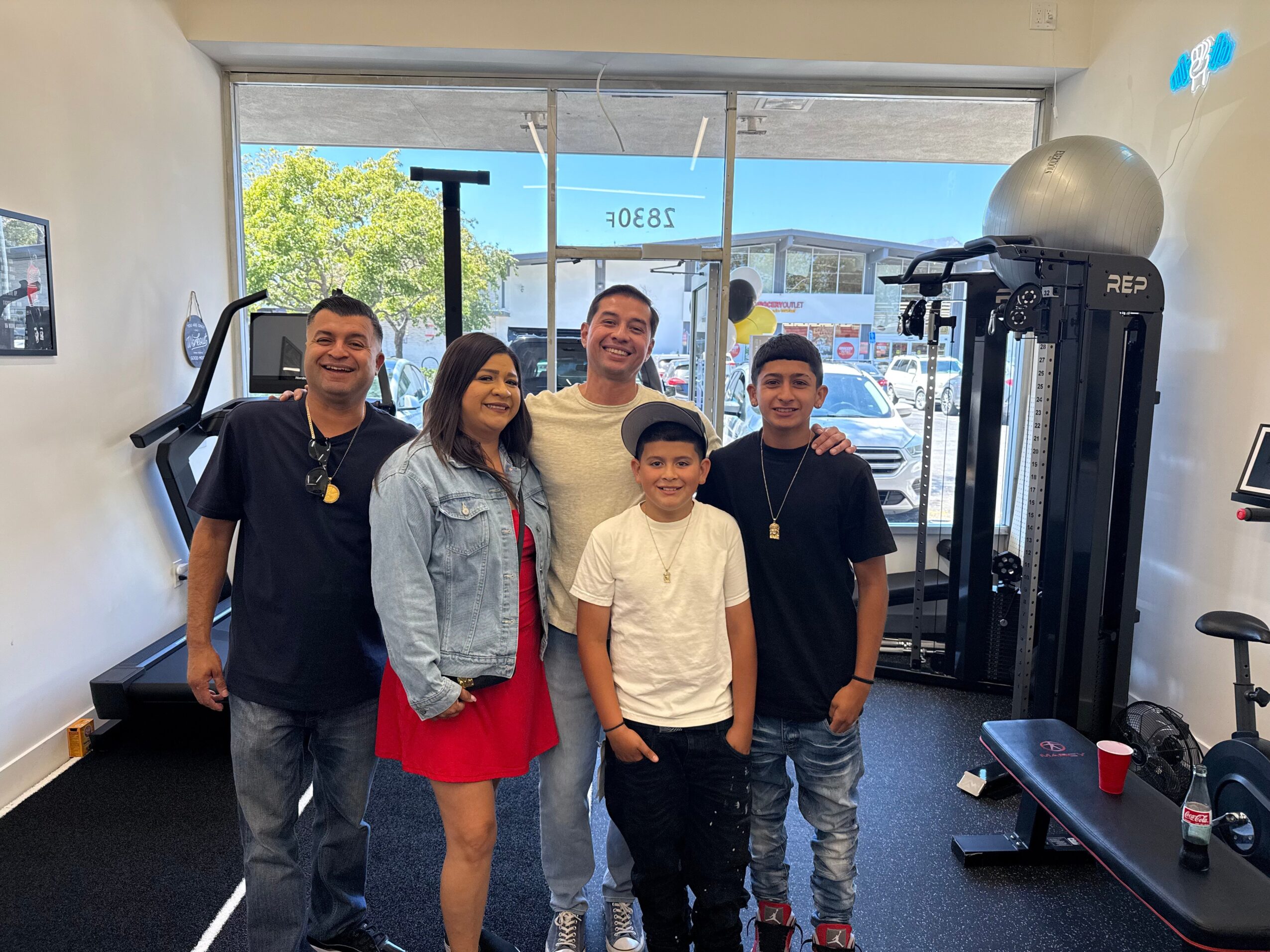 Group of five people smiling inside a gym, with exercise equipment and large window showing trees outside.