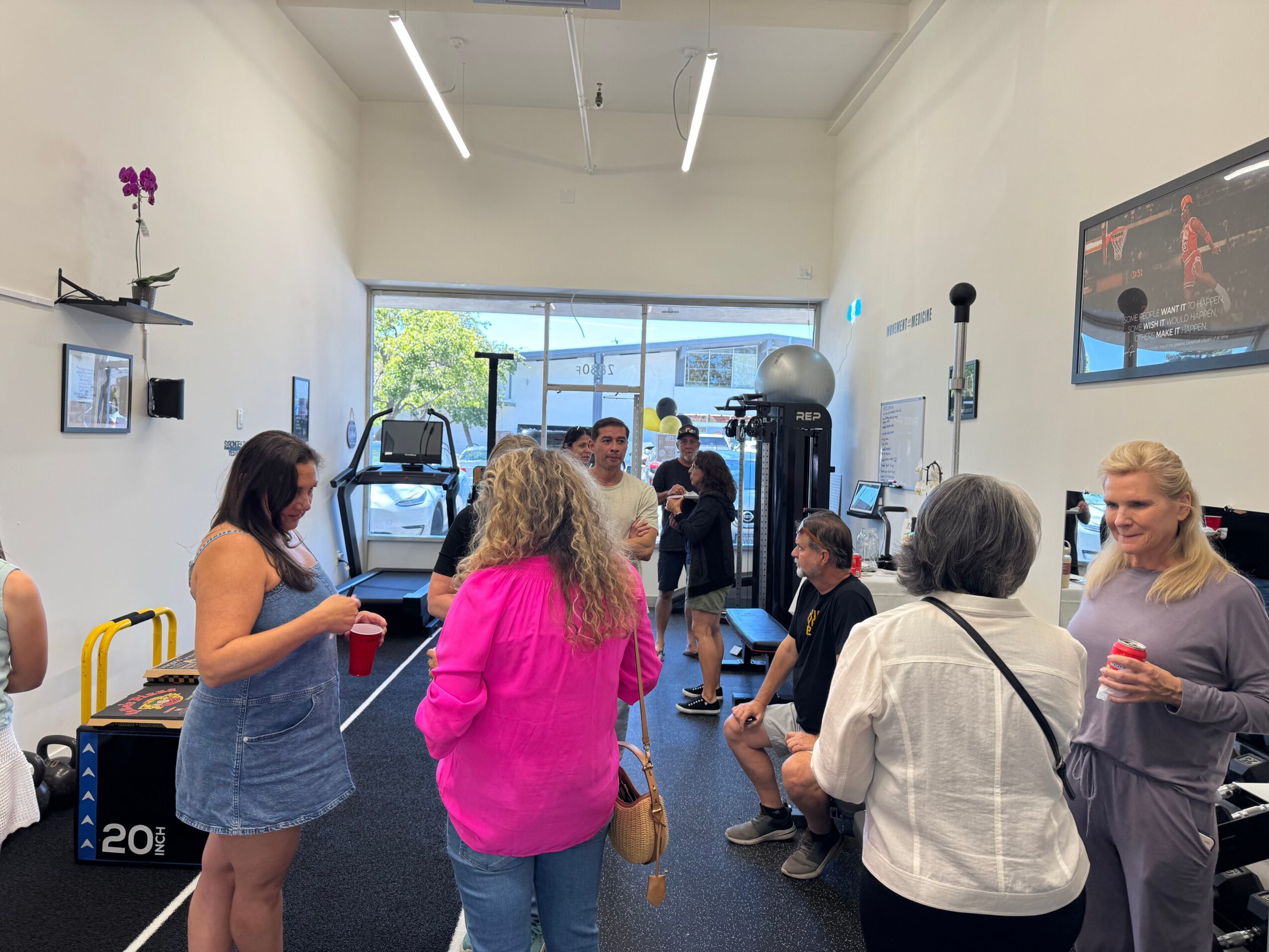 People in a gym with exercise equipment and a large window, some holding drinks, engaging in conversation.