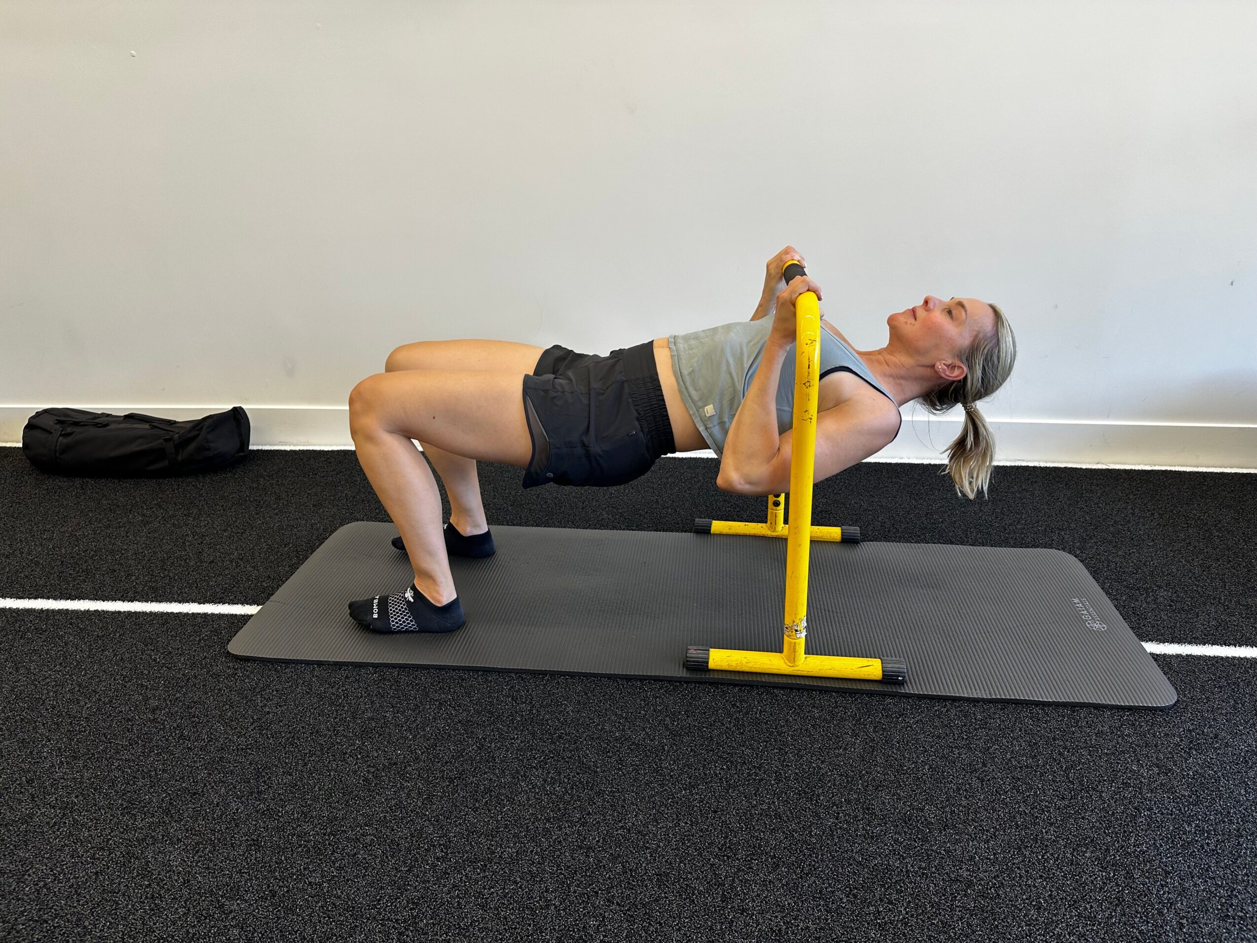 Woman lying on a bench with her head tilted back, performing a bridge exercise on a mat in a gym.