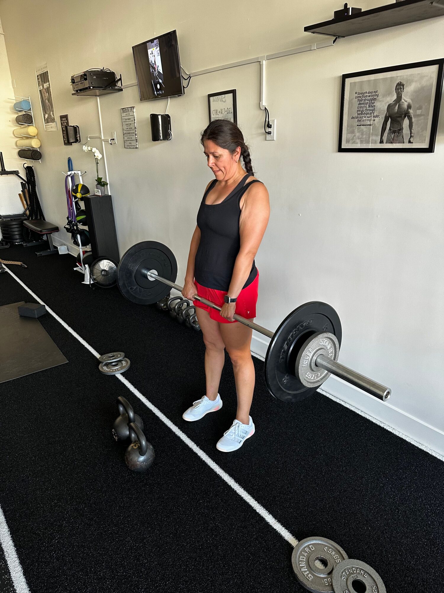 Woman in workout clothes standing in gym, holding a barbell, with weights and gym equipment around.