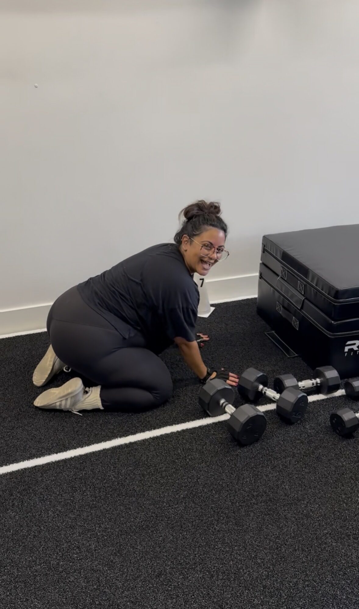 Person kneeling on gym floor, smiling, with dumbbells and exercise equipment nearby, against a plain wall.
