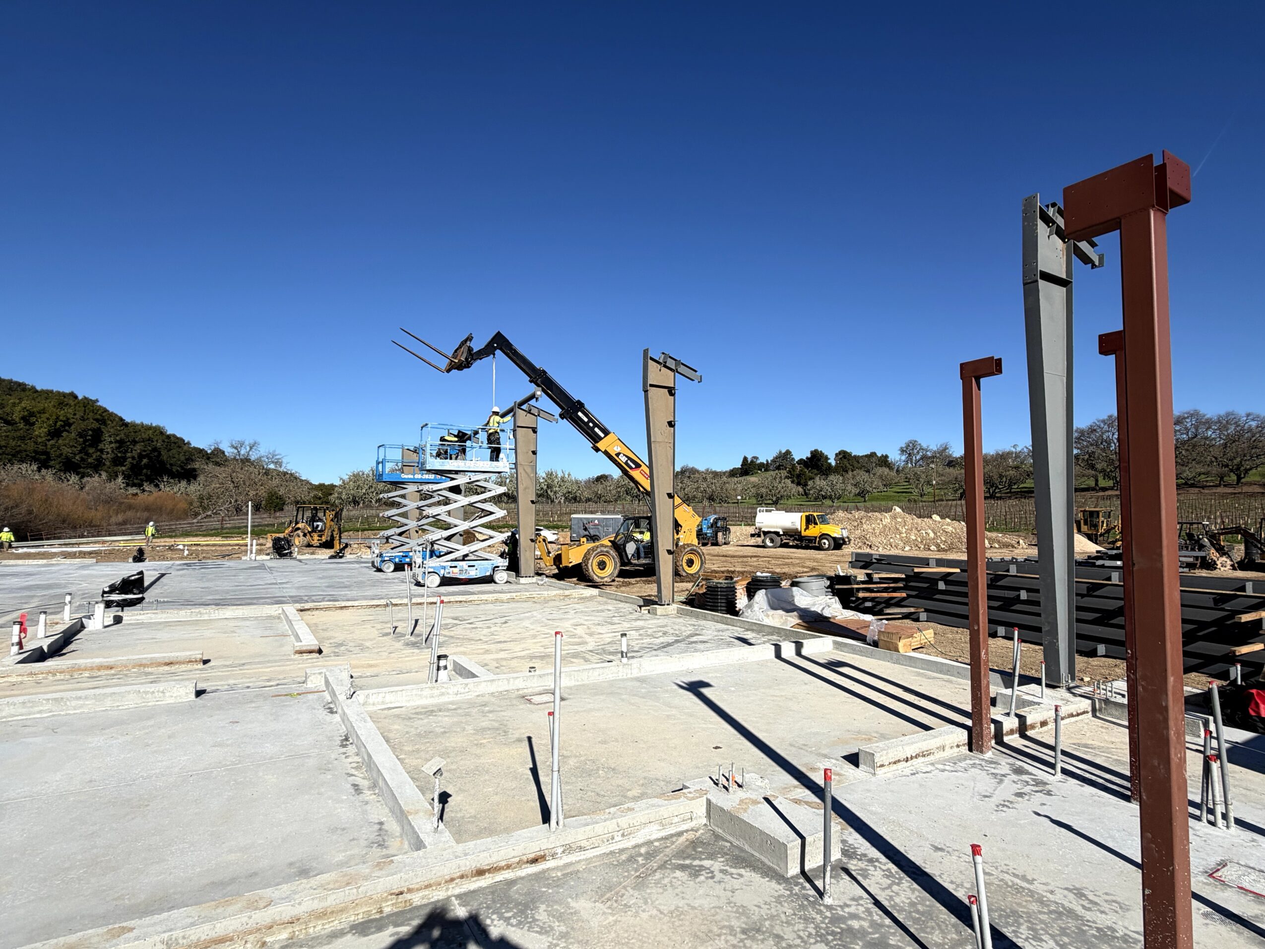 Construction site with machinery, steel beams, and tracks under a clear blue sky.