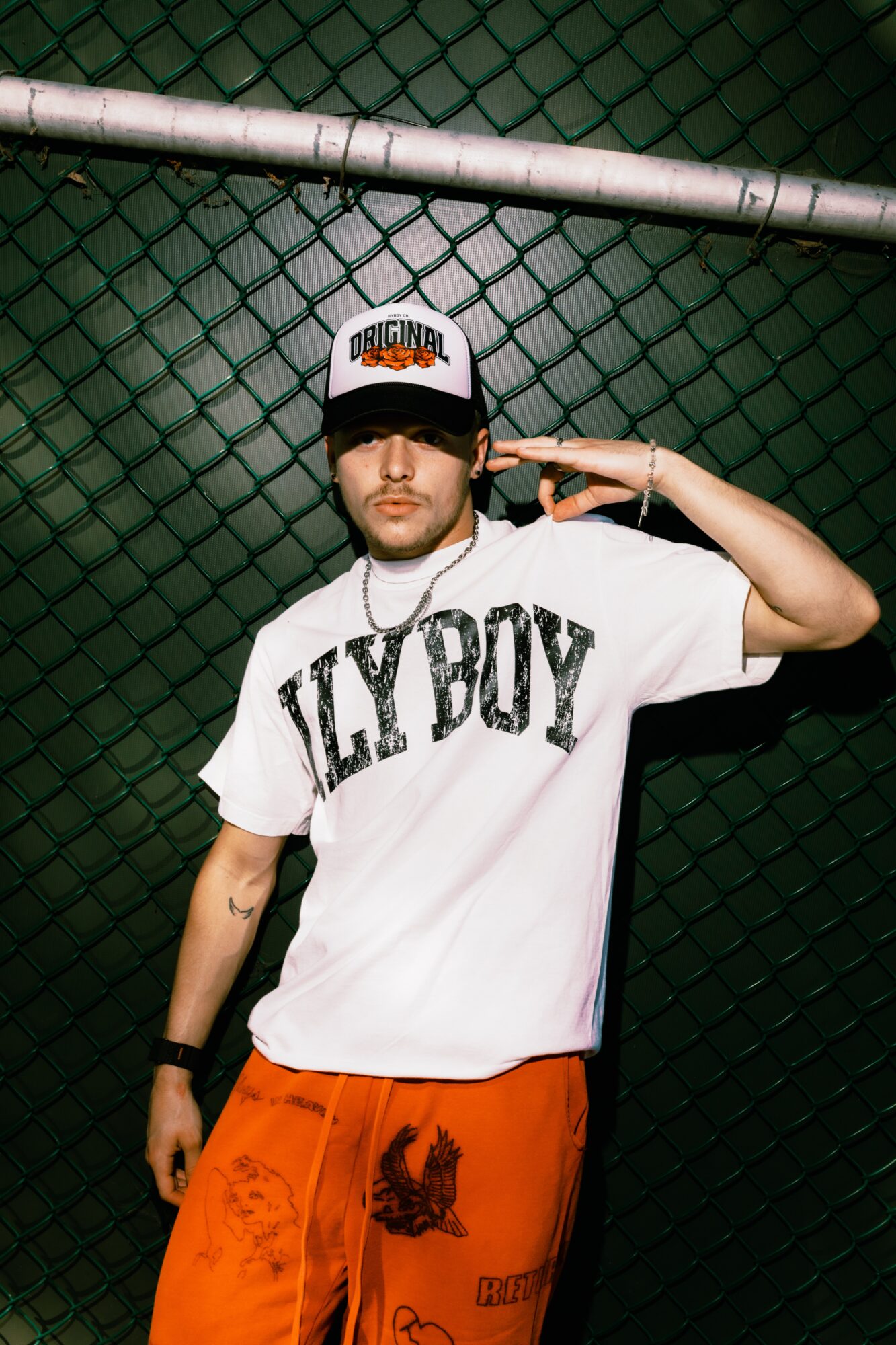 Person wearing a white T-shirt with 'FLY BOY' text, orange pants, and a cap, standing against a chain-link fence.