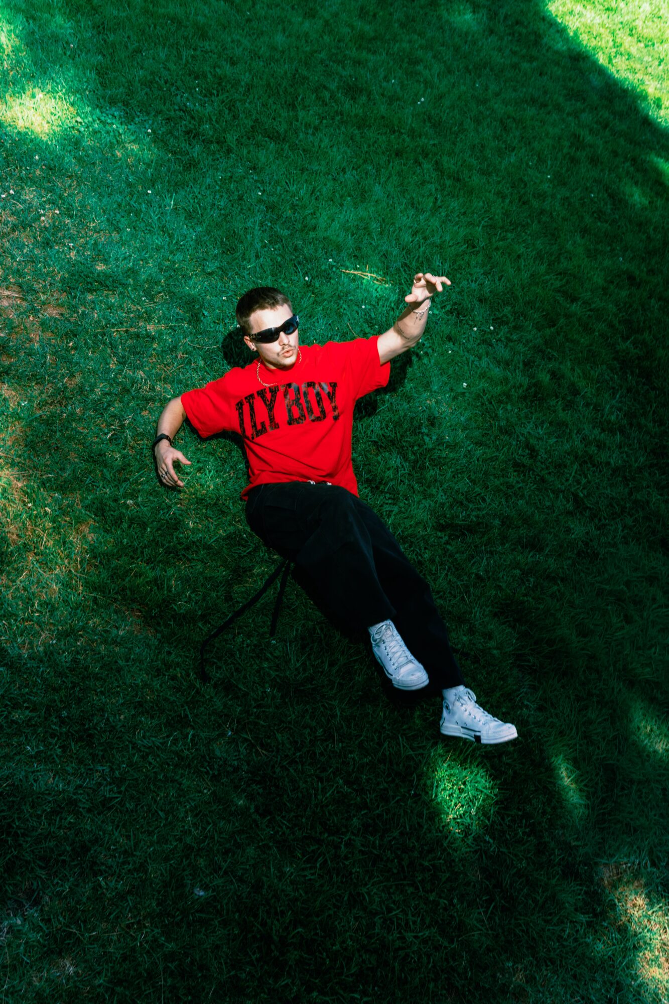 Person lying on grass wearing sunglasses, red shirt, black pants, and white shoes, with one arm raised.
