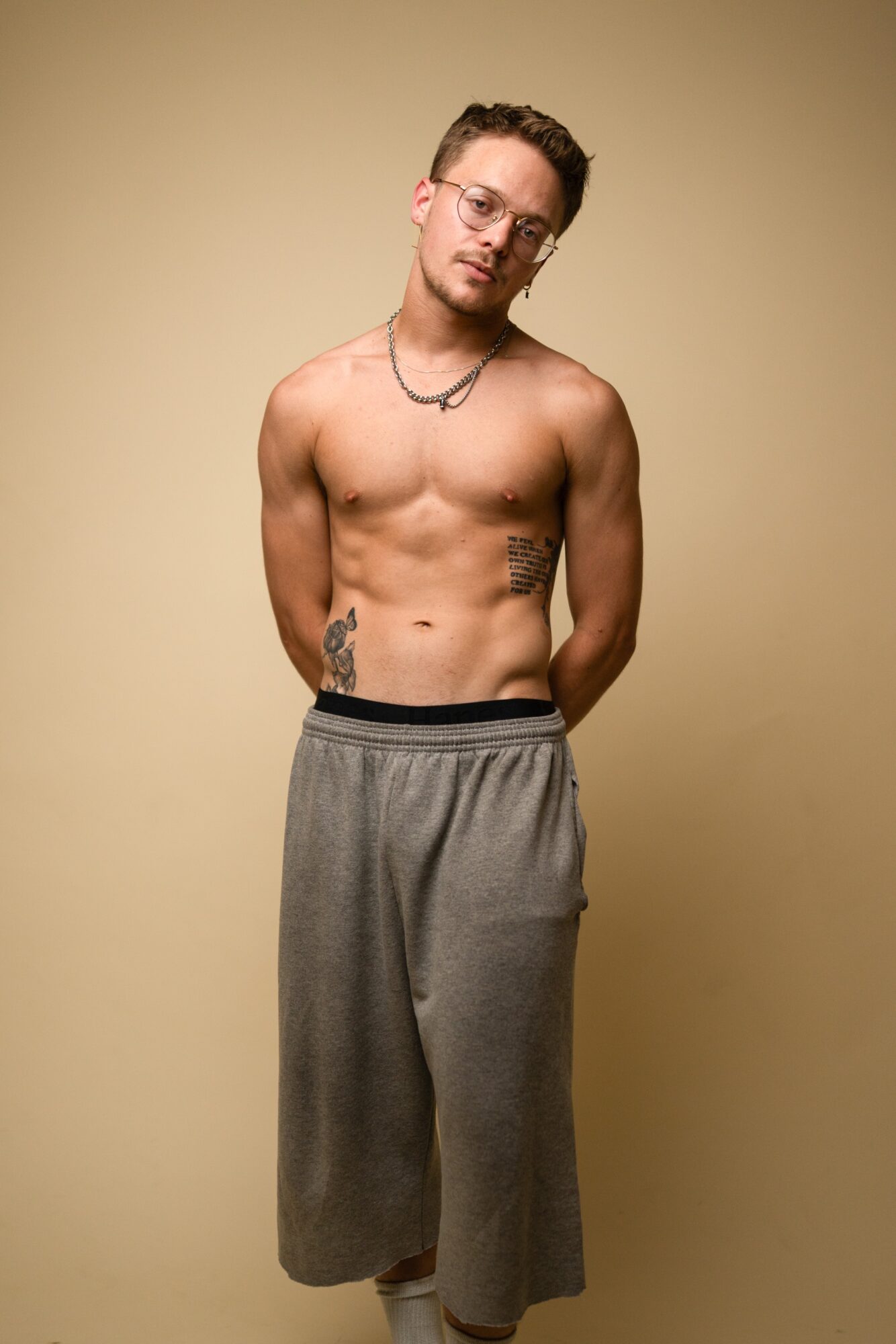 Young man with glasses, shirtless, wearing gray sweatpants, standing against a beige background.